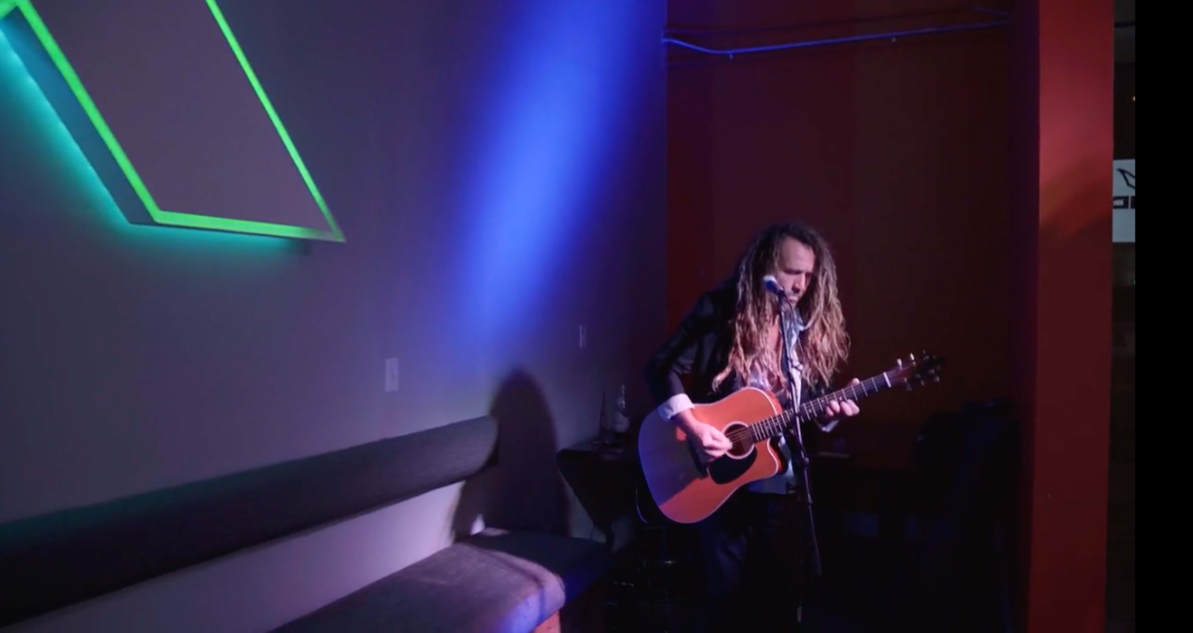A musician with long hair plays an acoustic guitar against a wall illuminated by colored lights.