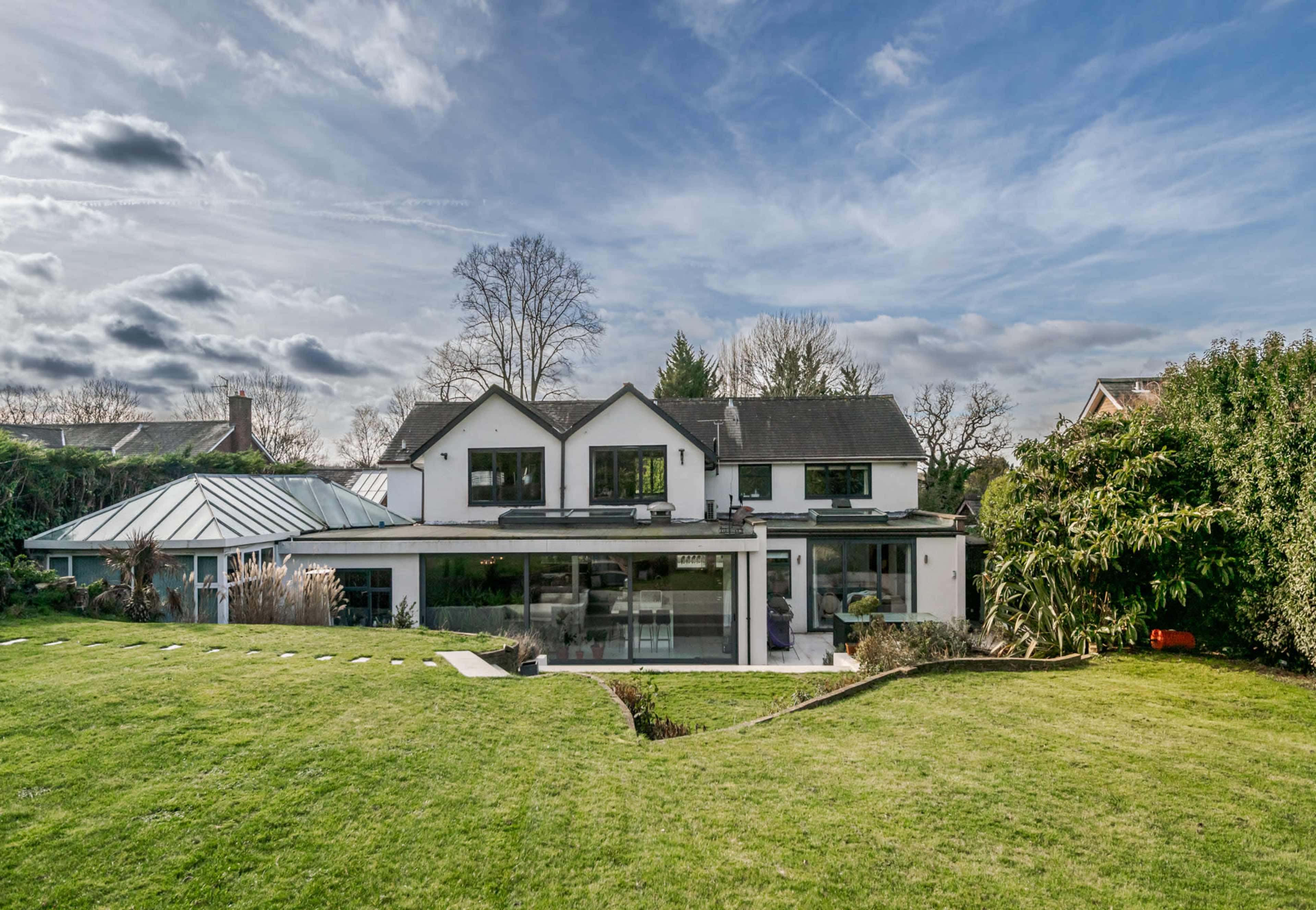 A modern house with a large glass extension, set in a spacious green lawn under a partly cloudy sky.