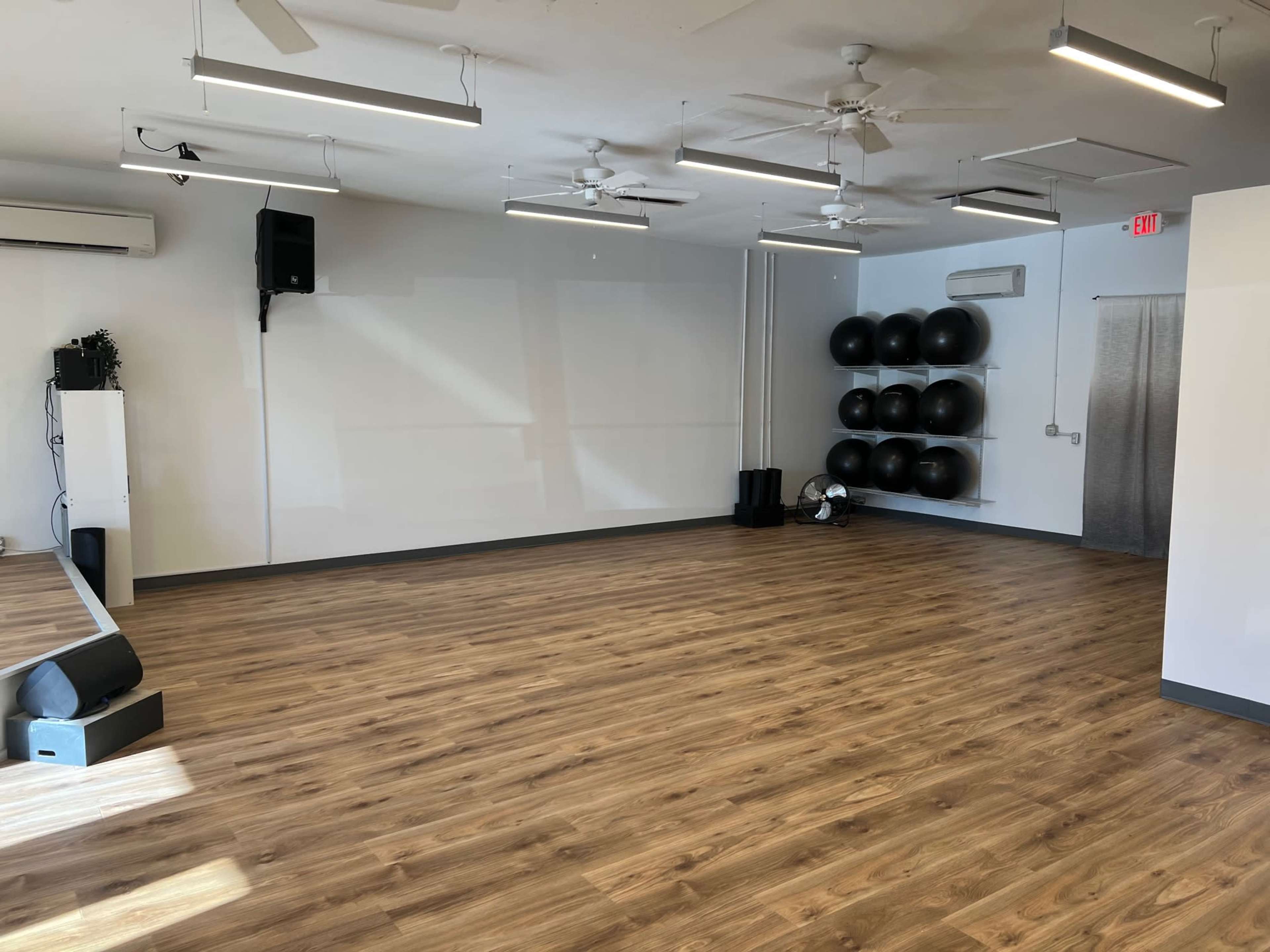 An empty fitness studio with wooden flooring, mirrors on the wall, and exercise balls stacked in the corner.