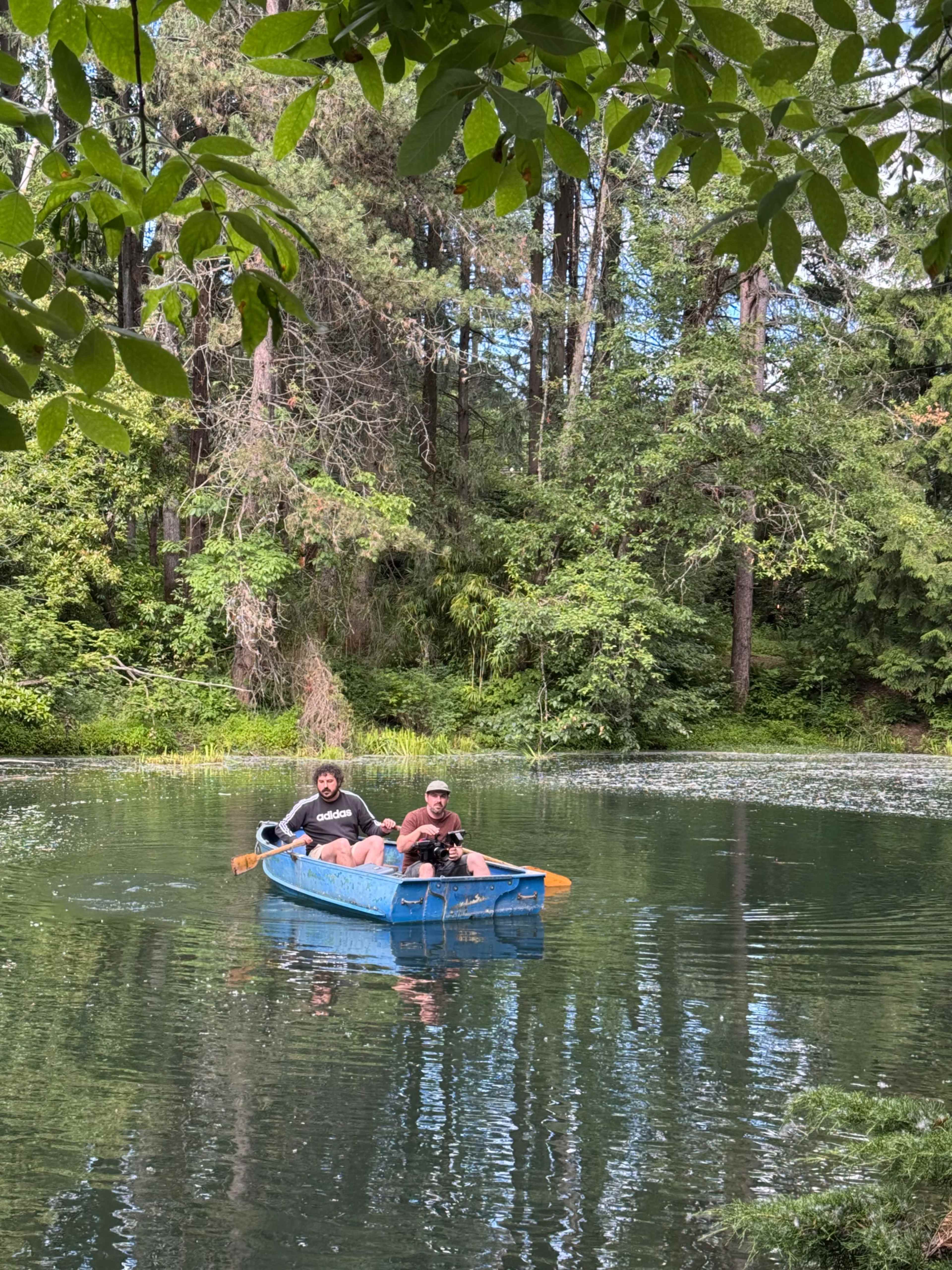 Two men paddle a blue boat on a calm, green lake surrounded by trees.