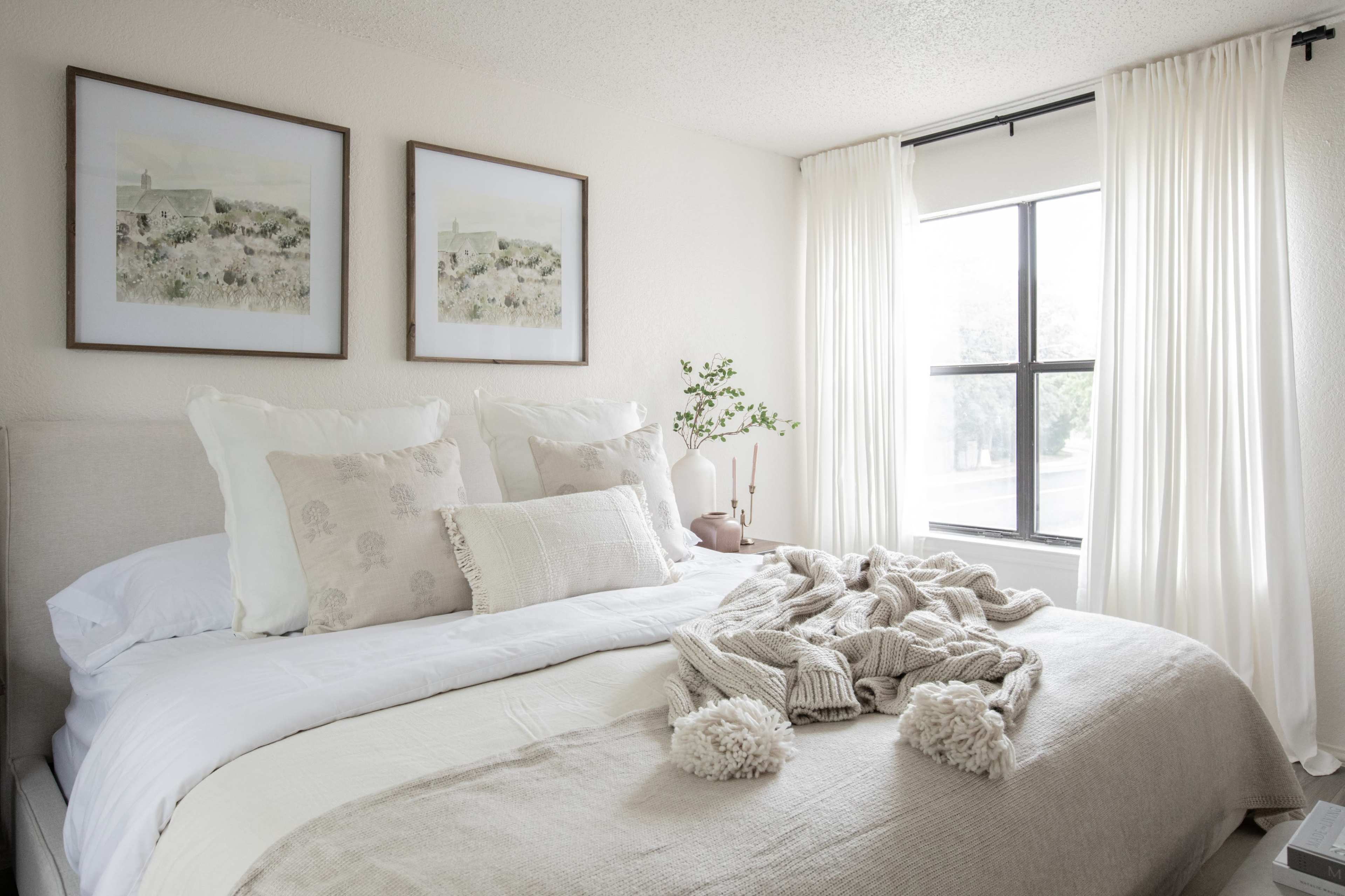 A neatly arranged bedroom with a large bed, adorned with light-colored pillows and a cozy throw, accompanied by two framed artworks on the wall and soft white curtains hanging by the window.