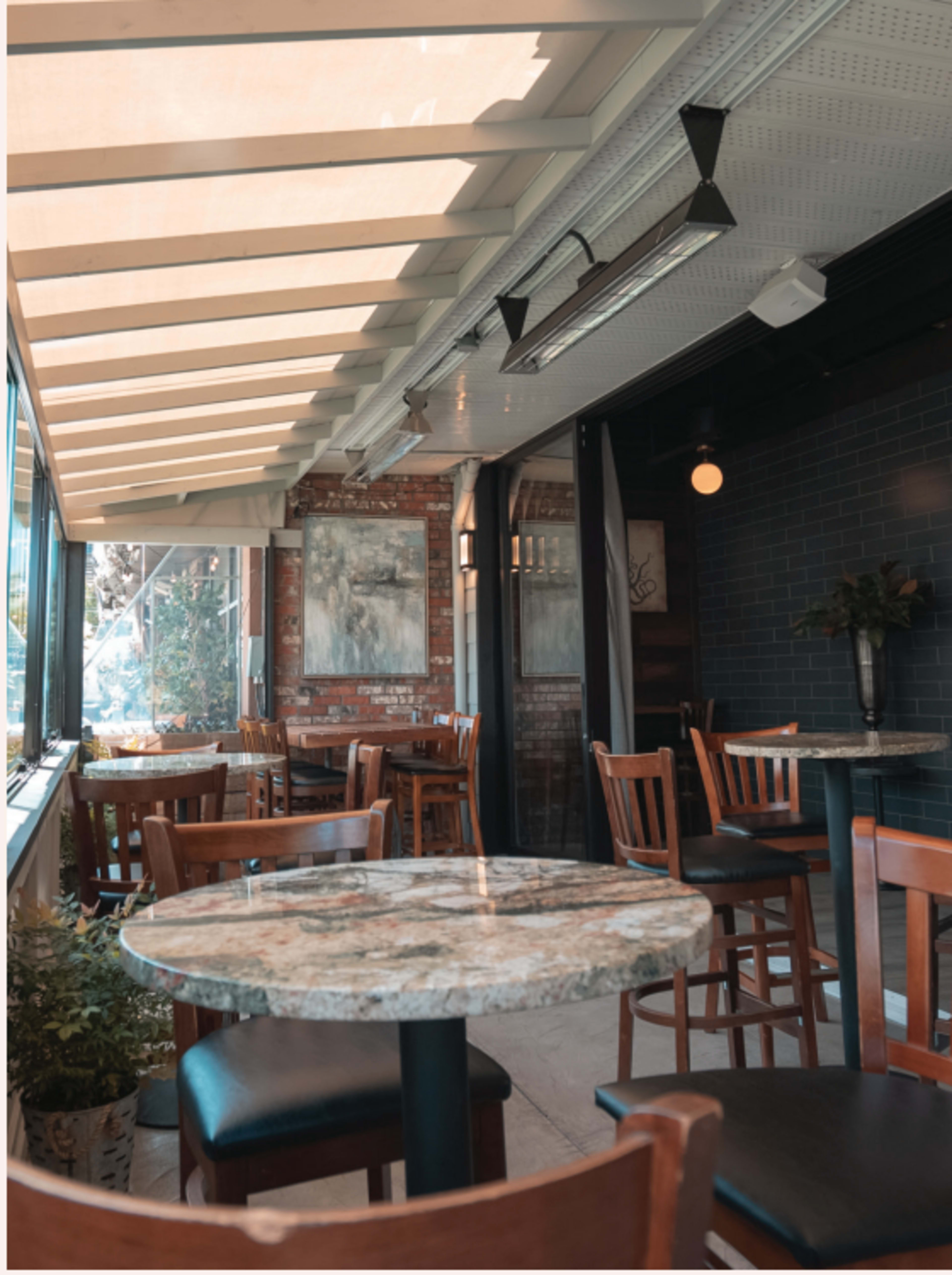 A bright, inviting café with wooden tables and chairs arranged near large windows and exposed brick walls.