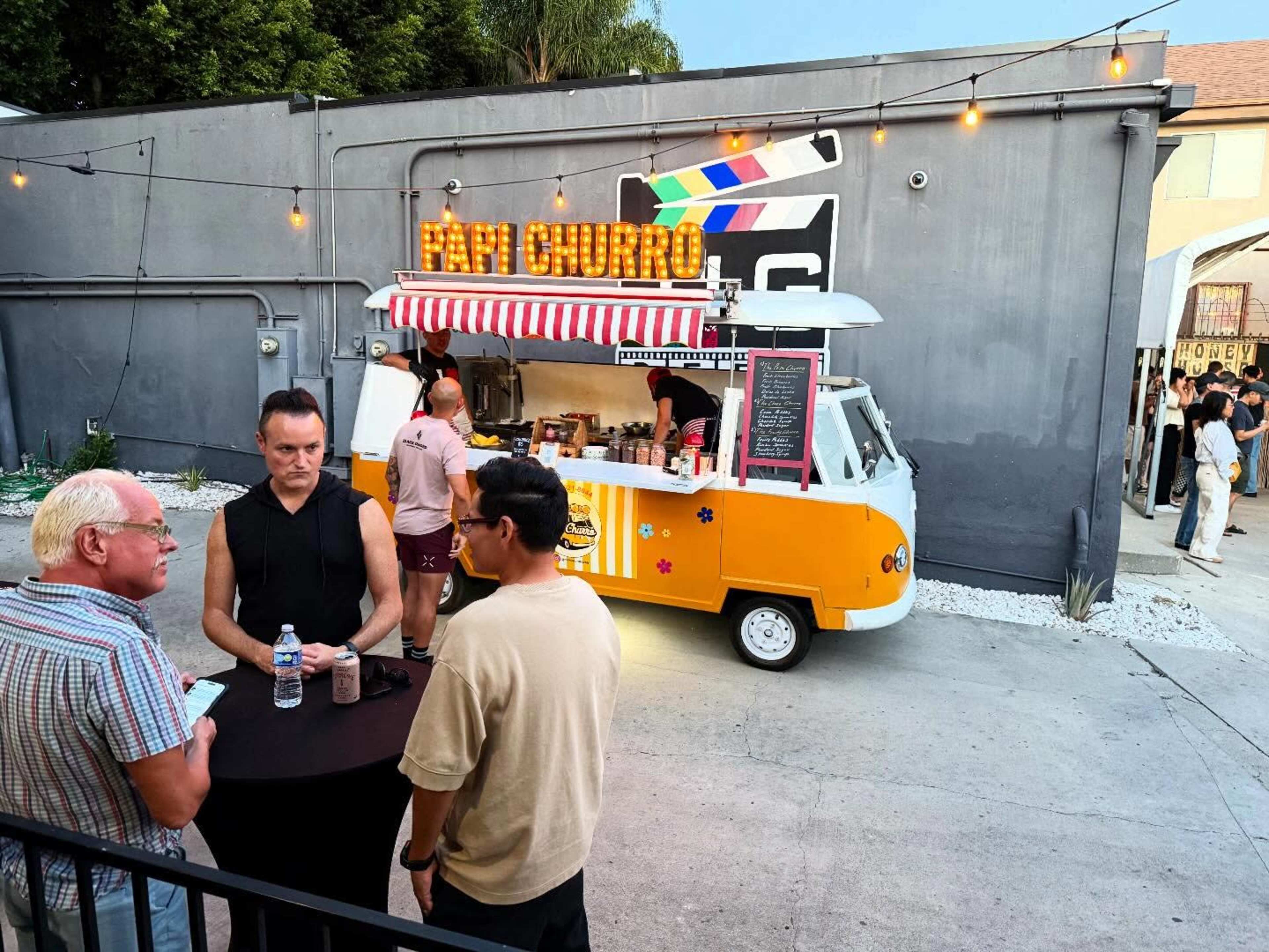 A food truck named "Mini Churro" is parked next to a gathering of people at an outdoor event, with string lights overhead and a gray wall in the background.