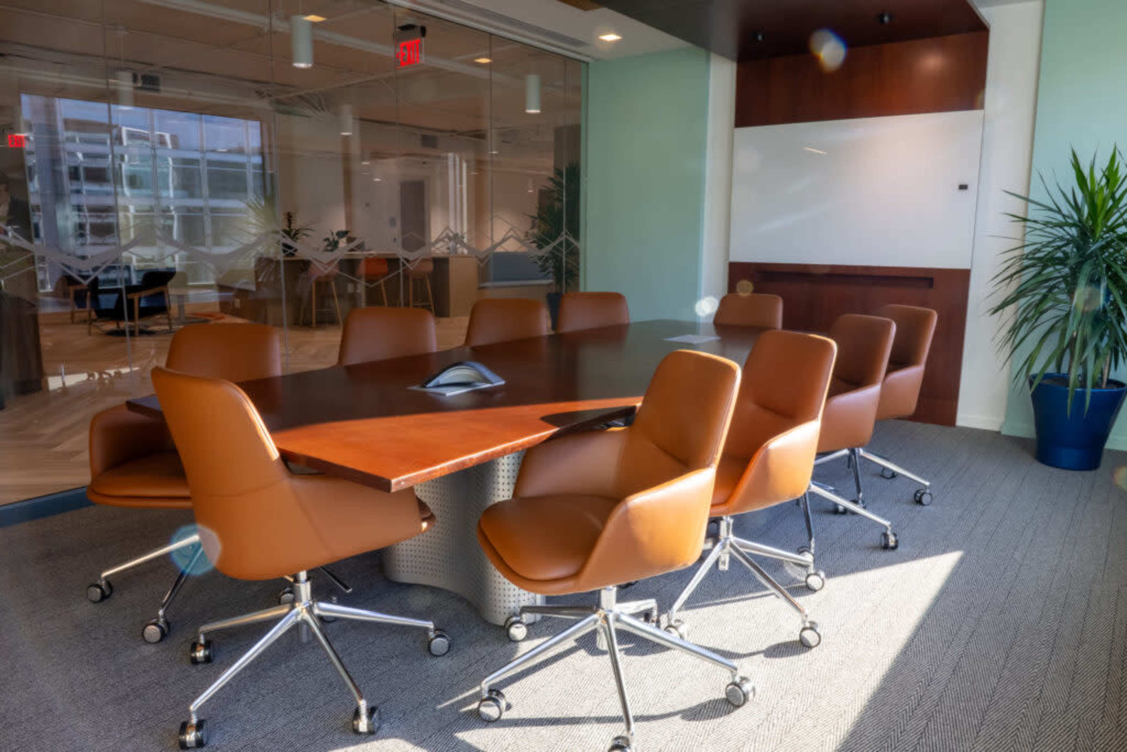 A modern conference room features a long wooden table surrounded by eight brown leather chairs, with a large window letting in natural light.