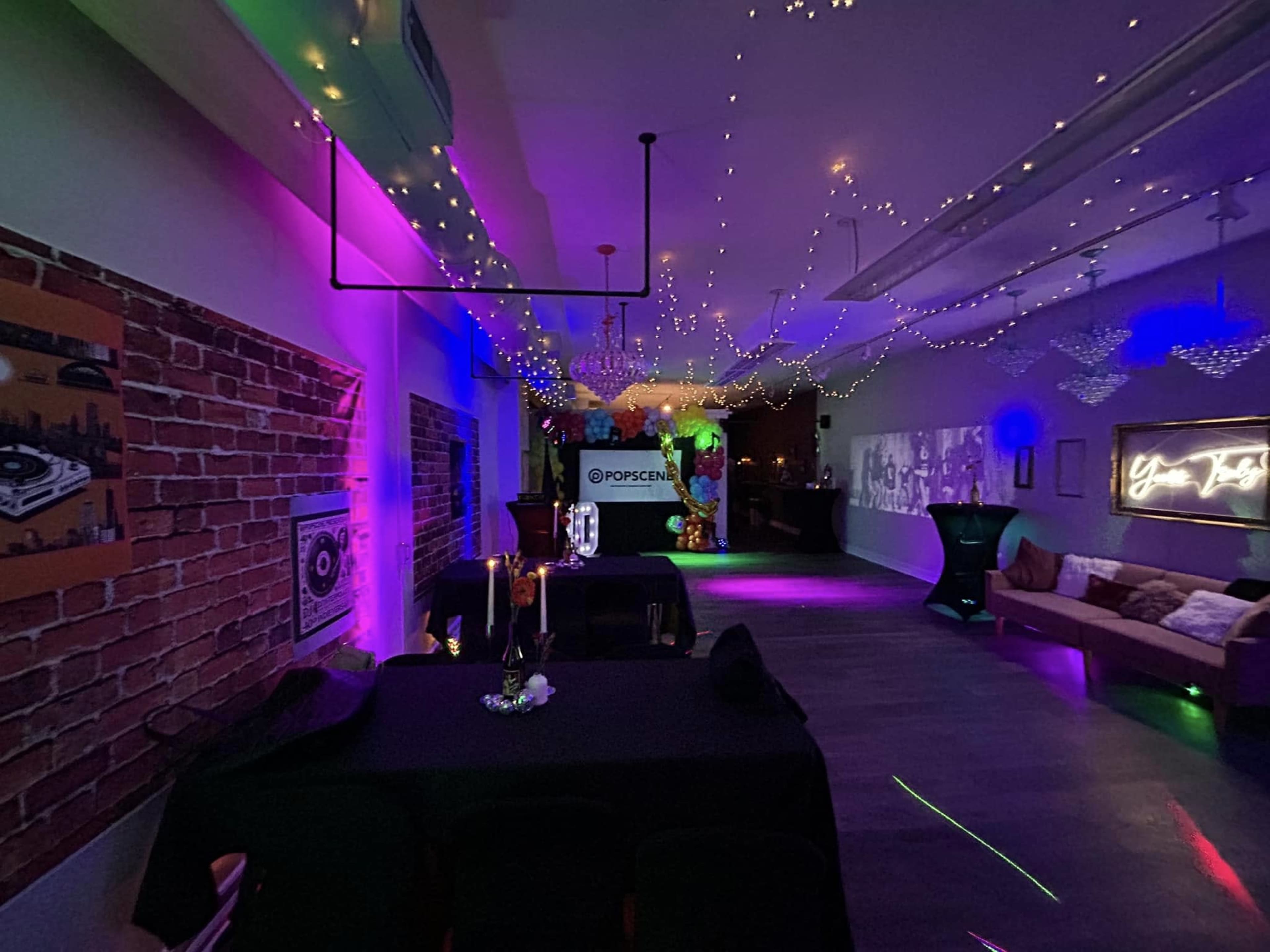 The image shows a dimly lit event space decorated with string lights and neon colors, featuring tables and a large projection screen.