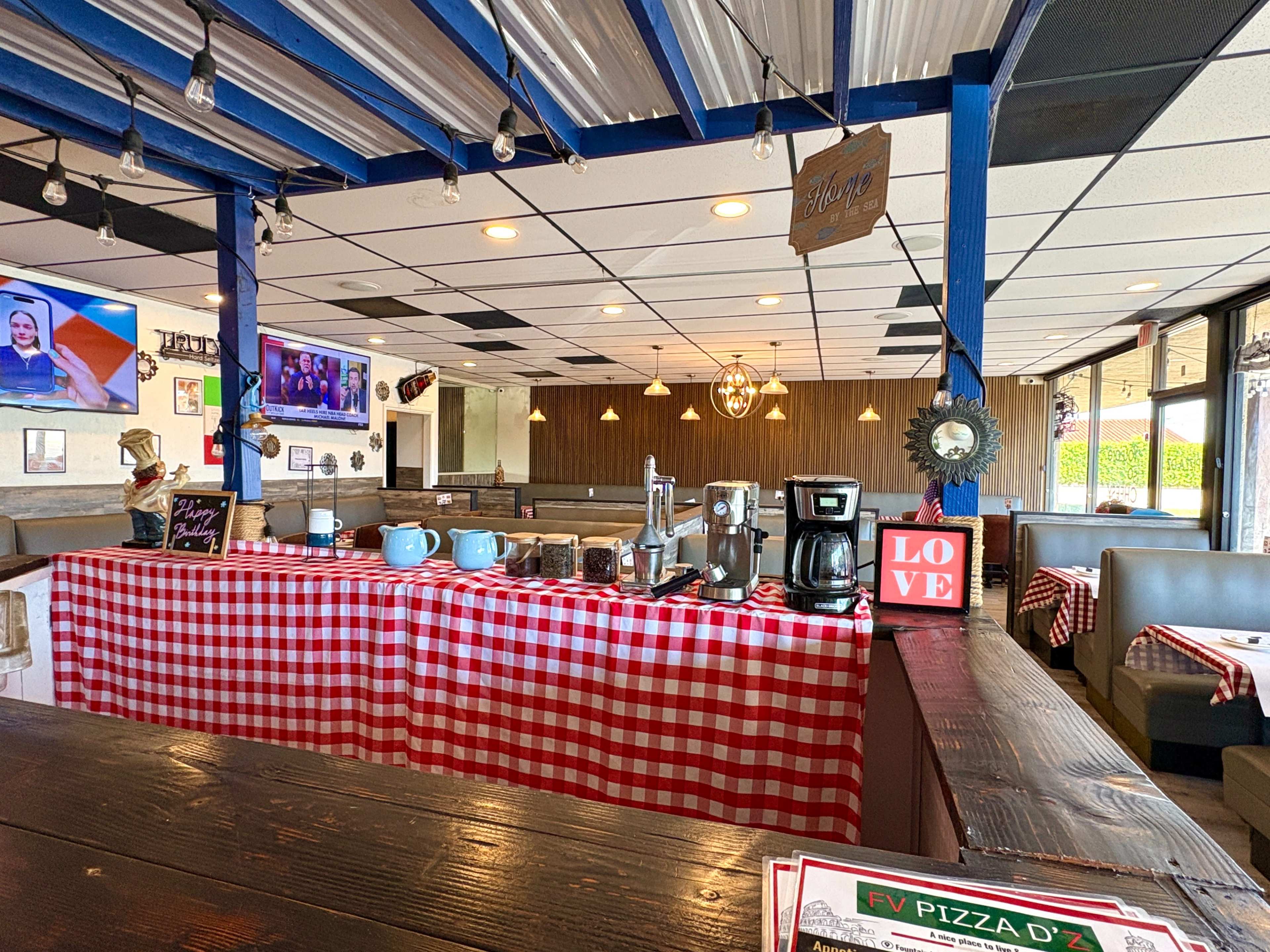 The image shows the interior of a restaurant with red and white checkered tablecloths, a counter showcasing coffee equipment, and a television displaying images on the wall.