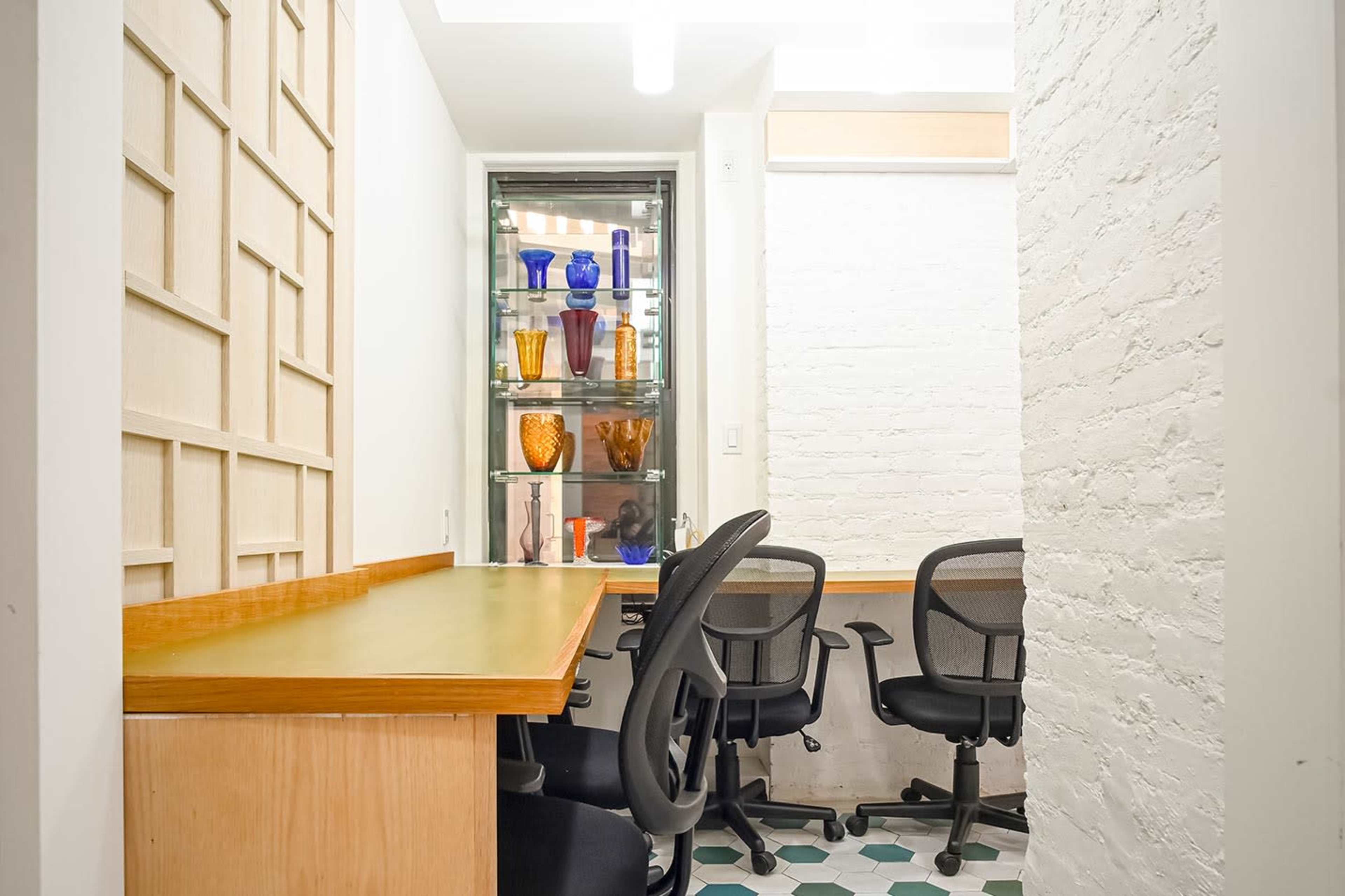 The image shows a small office space featuring a wooden countertop, several office chairs, and a glass display case filled with colorful vases.