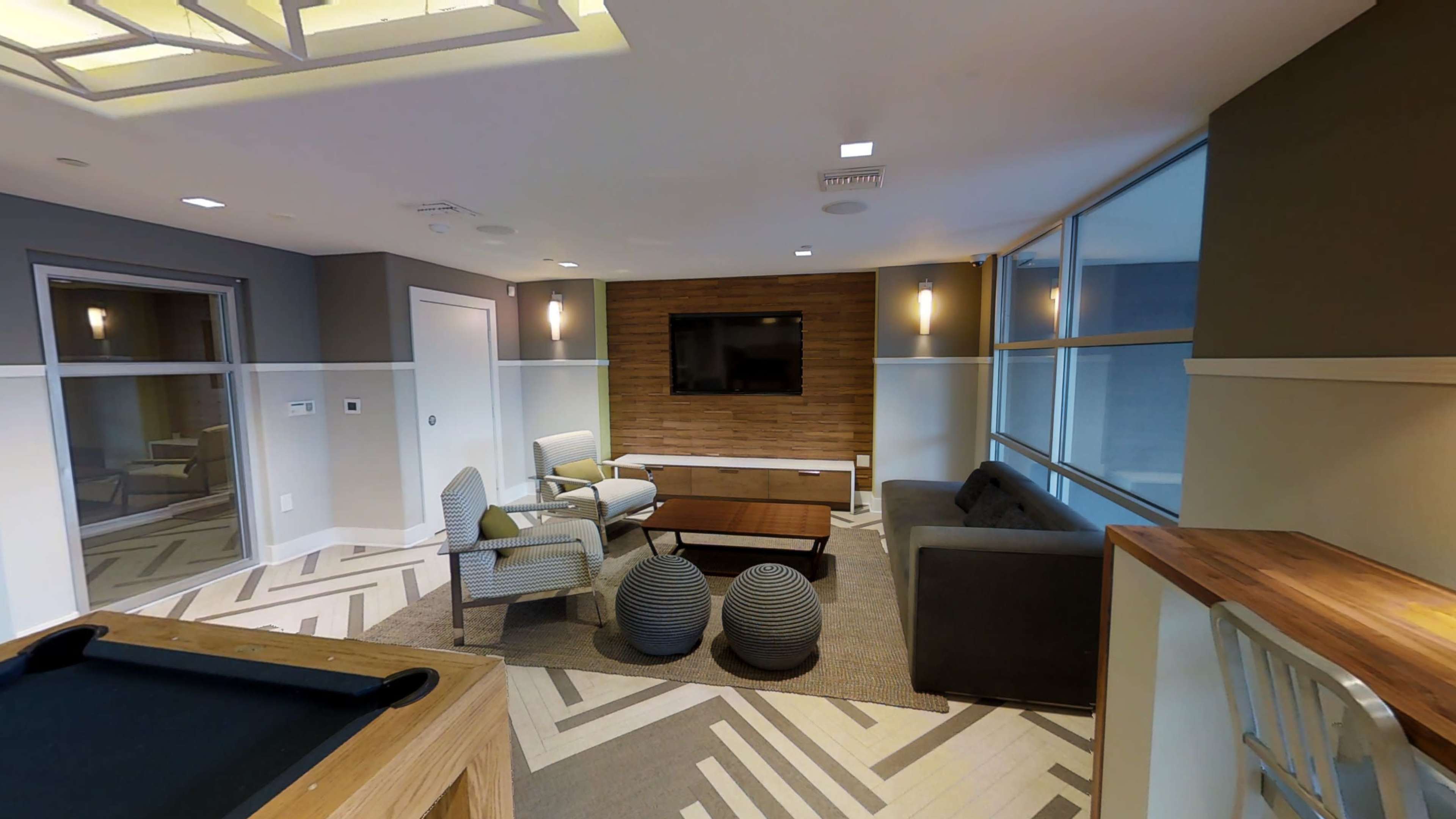 A modern lounge area with a flat-screen TV, a coffee table, seating arrangement, and decorative elements like patterned rugs and large windows.