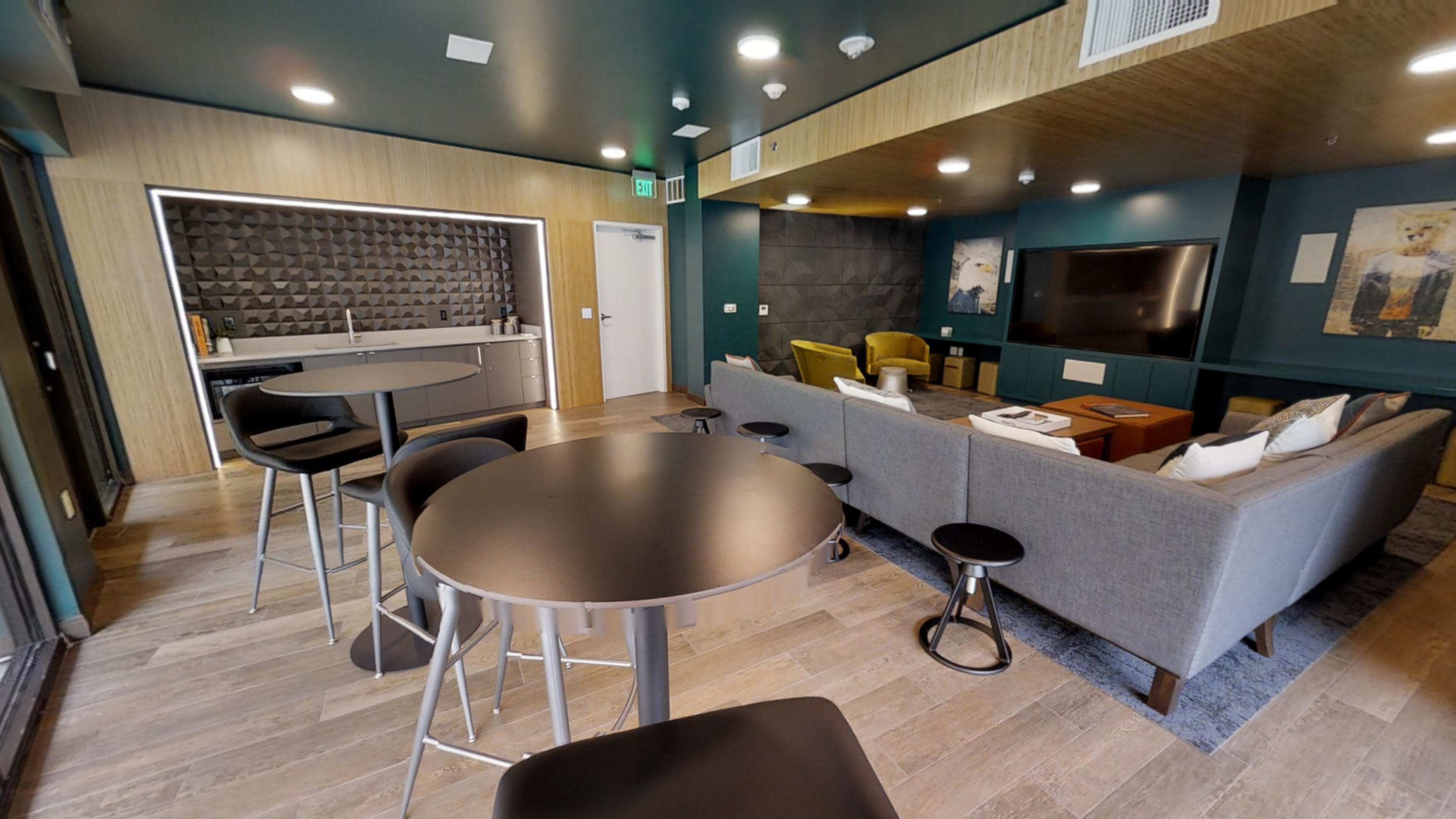 A modern lounge area with seating, a television, and a kitchenette featuring bar stools and tables.