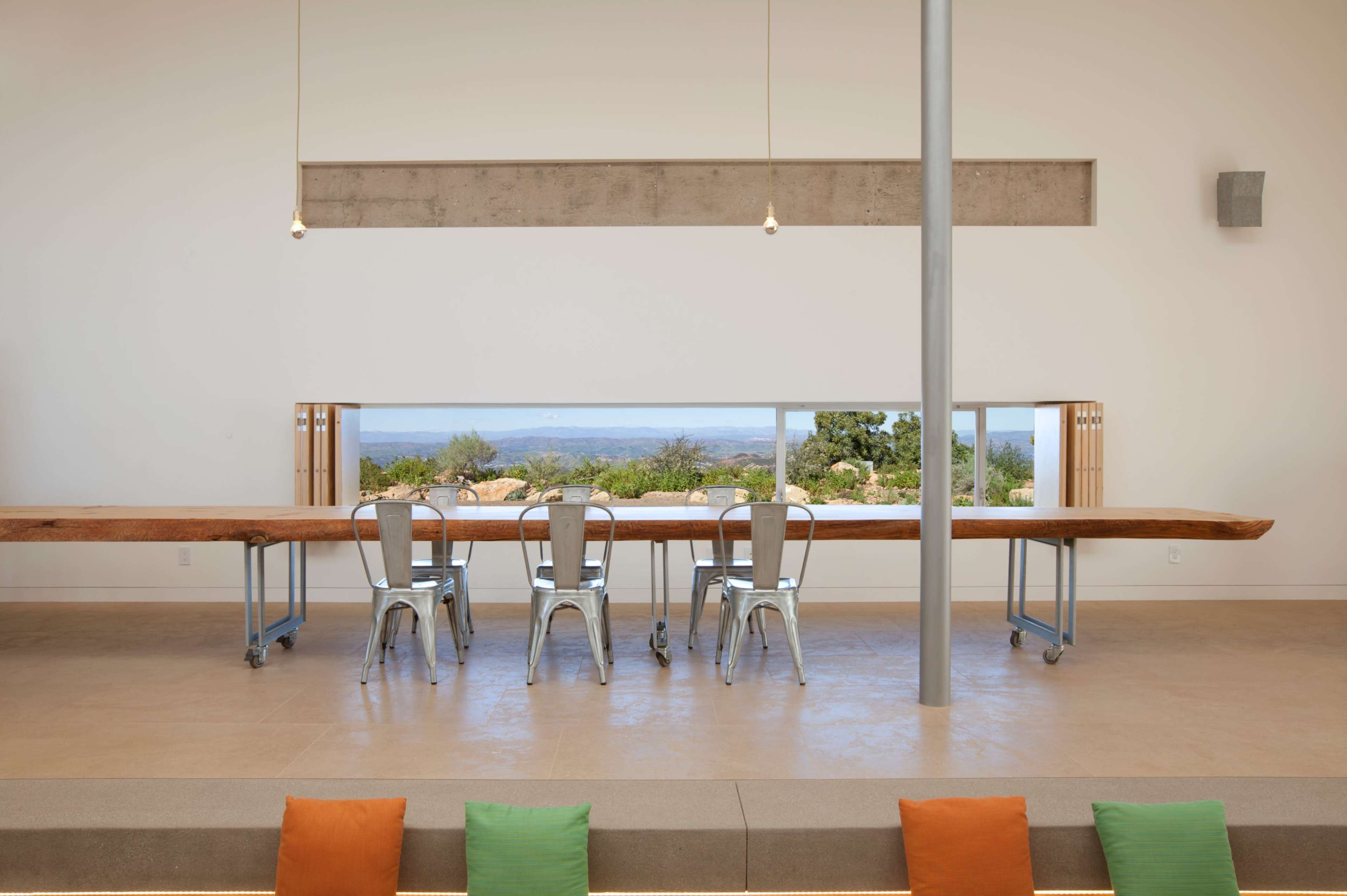 A long wooden table with metal chairs is situated in a minimalist room featuring a large window that frames a view of the landscape beyond.