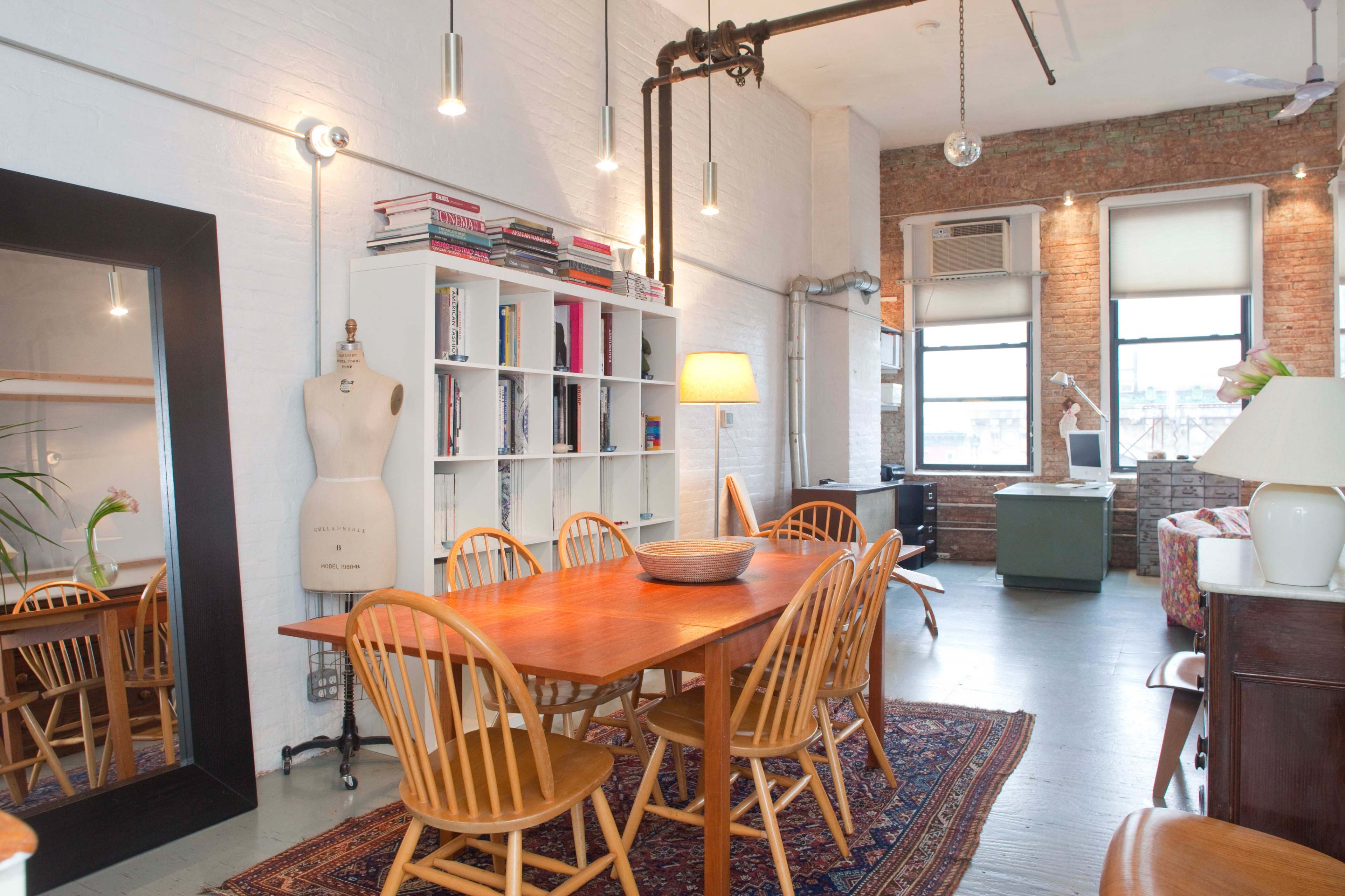 An airy loft space with a dining table surrounded by wooden chairs, a bookshelf, and large windows revealing a brick wall.