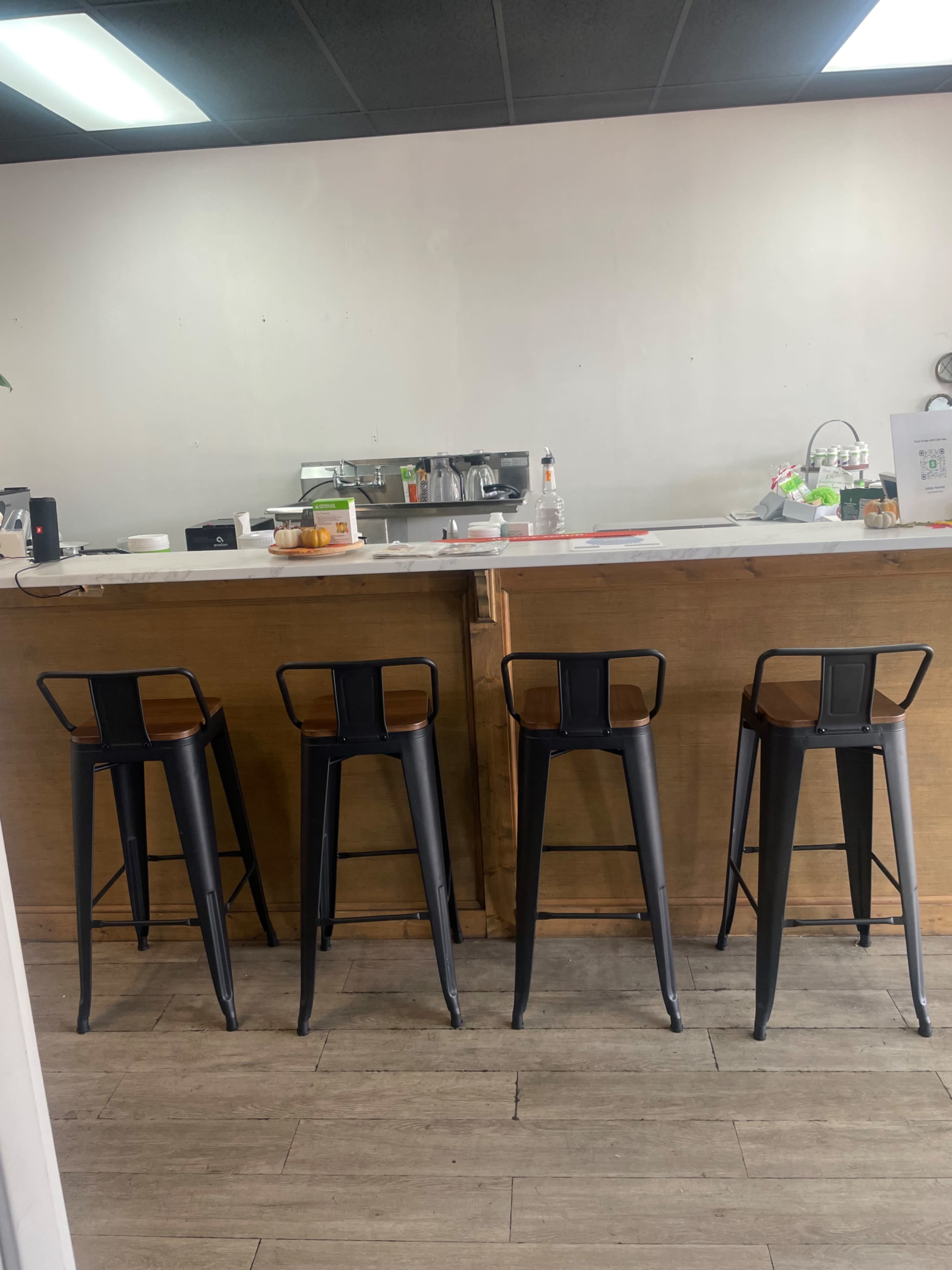 The image shows a row of four black metal stools lined up in front of a wooden counter in a minimalist café setting.