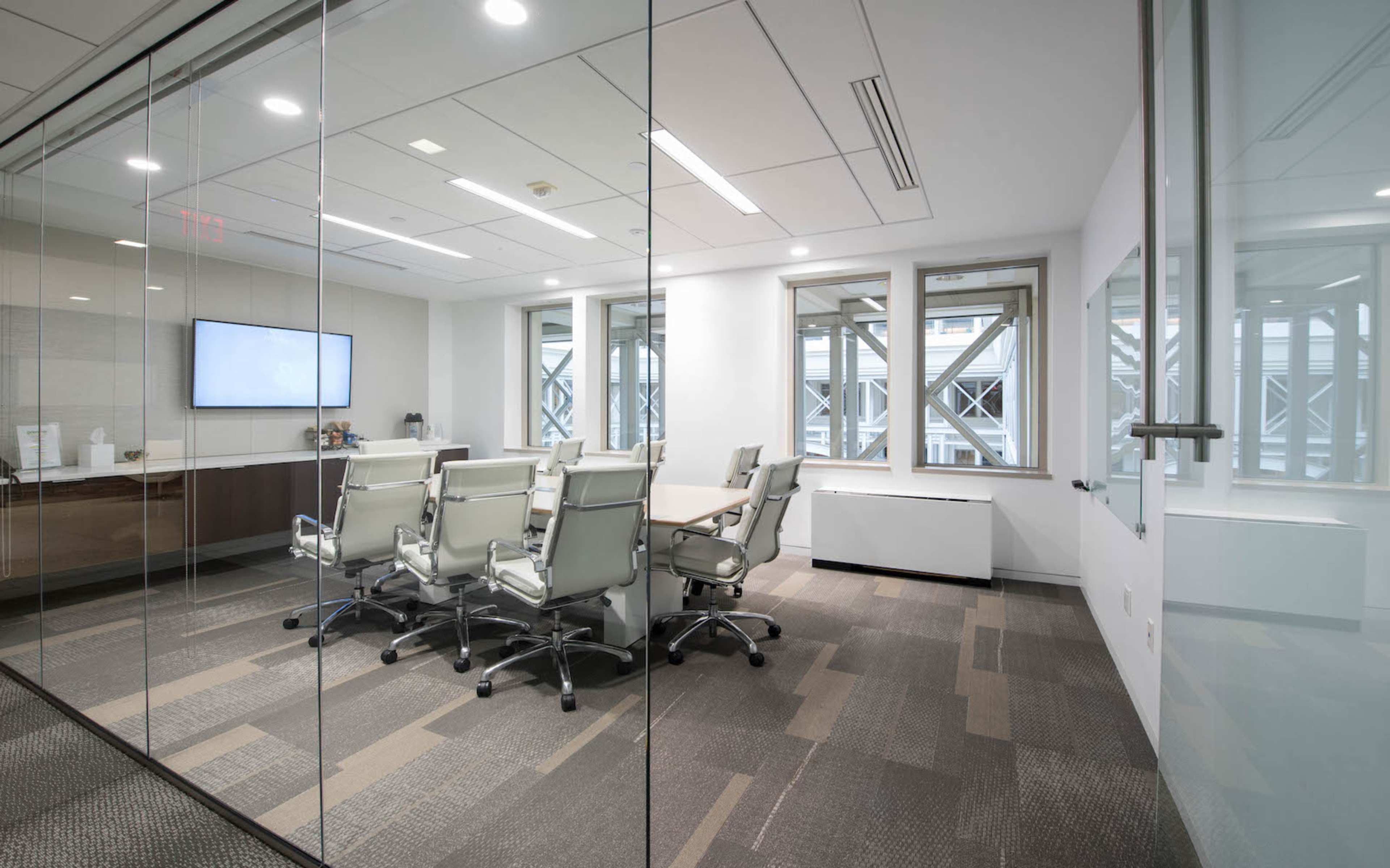 A modern conference room with a large table surrounded by several white chairs, glass walls, and a screen mounted on the wall.