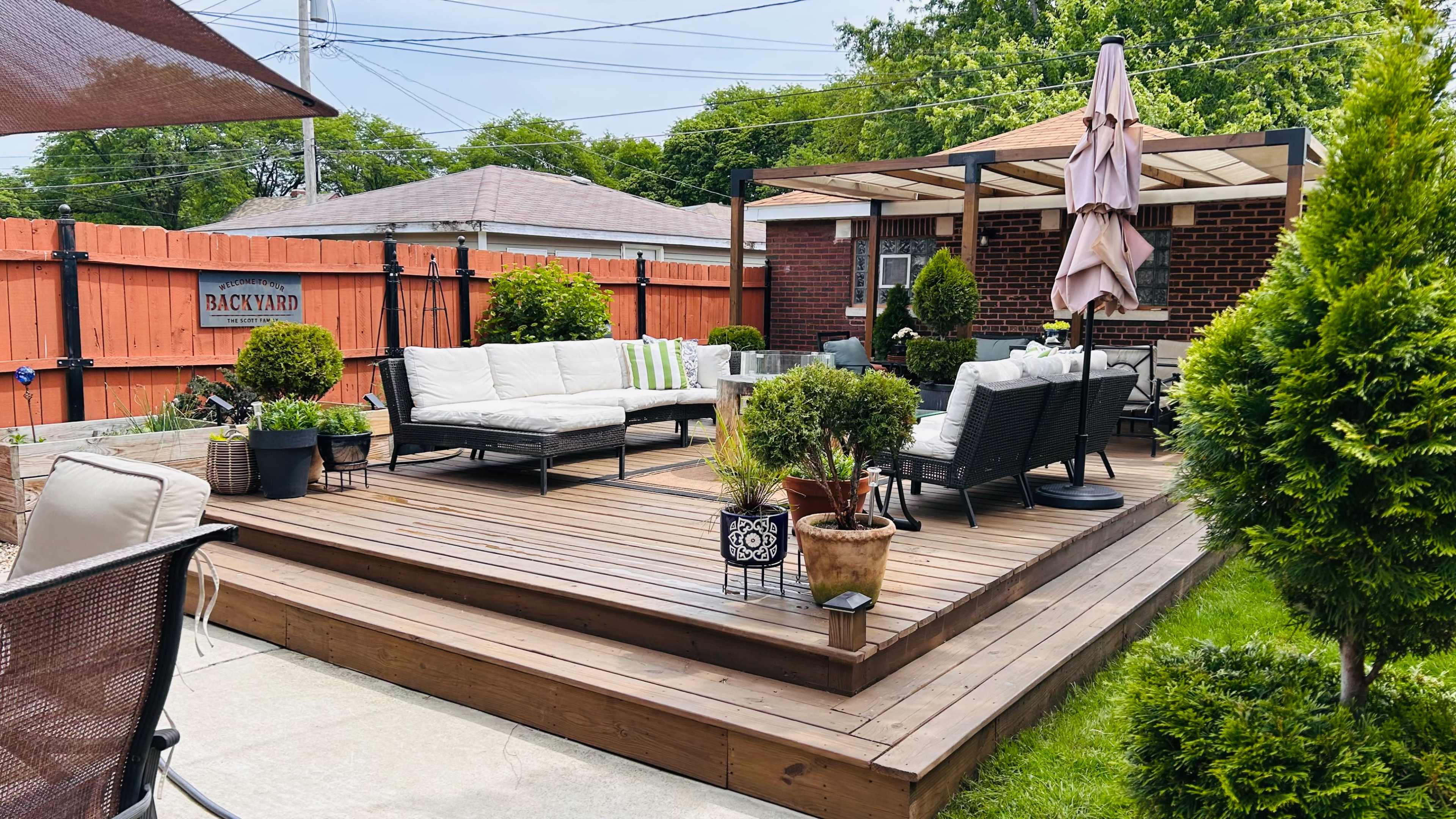 The image shows a wooden deck furnished with seating, surrounded by potted plants and a patio umbrella, in a landscaped backyard.