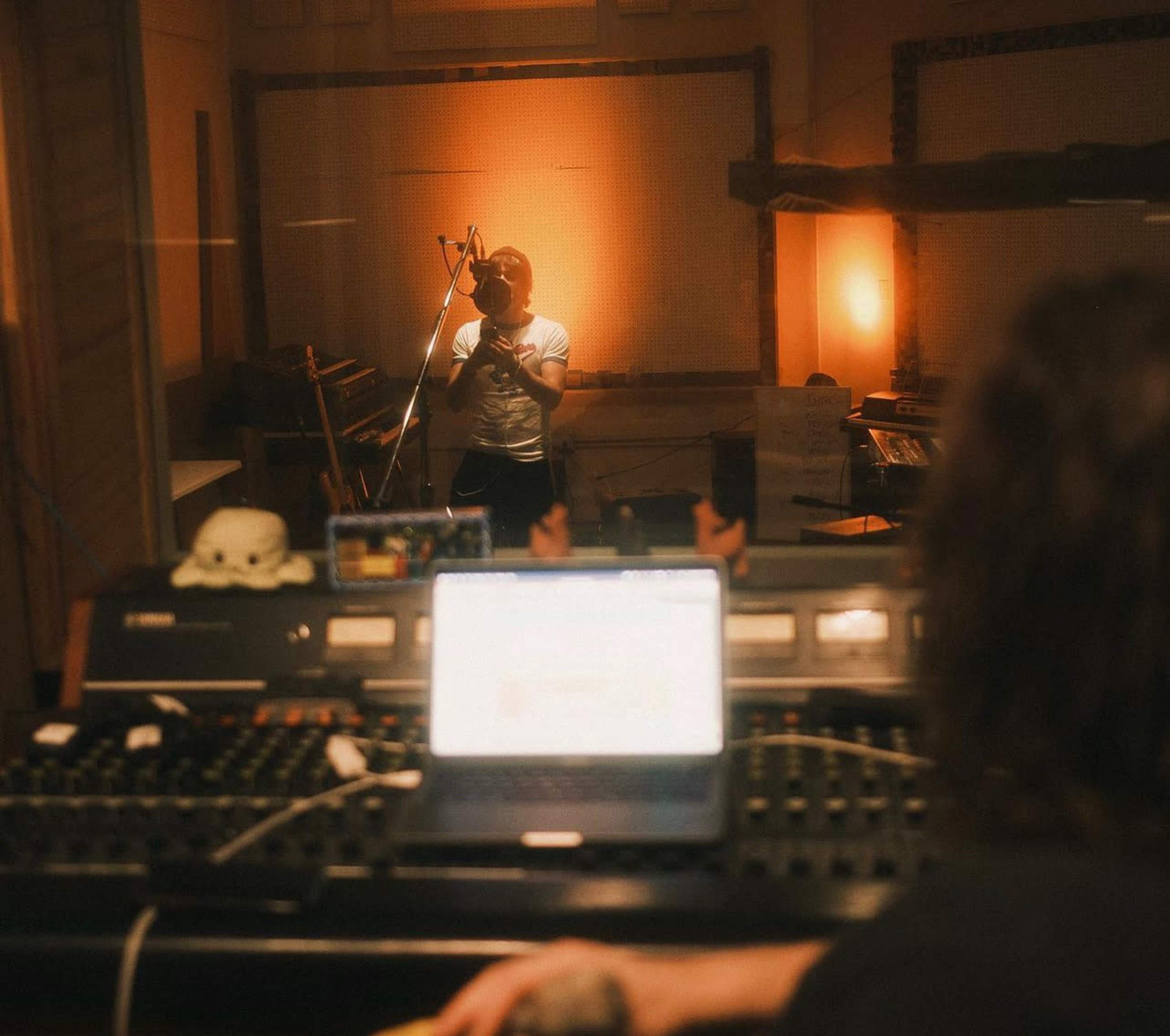 A person stands in a recording studio while another operates a mixing console in the foreground.