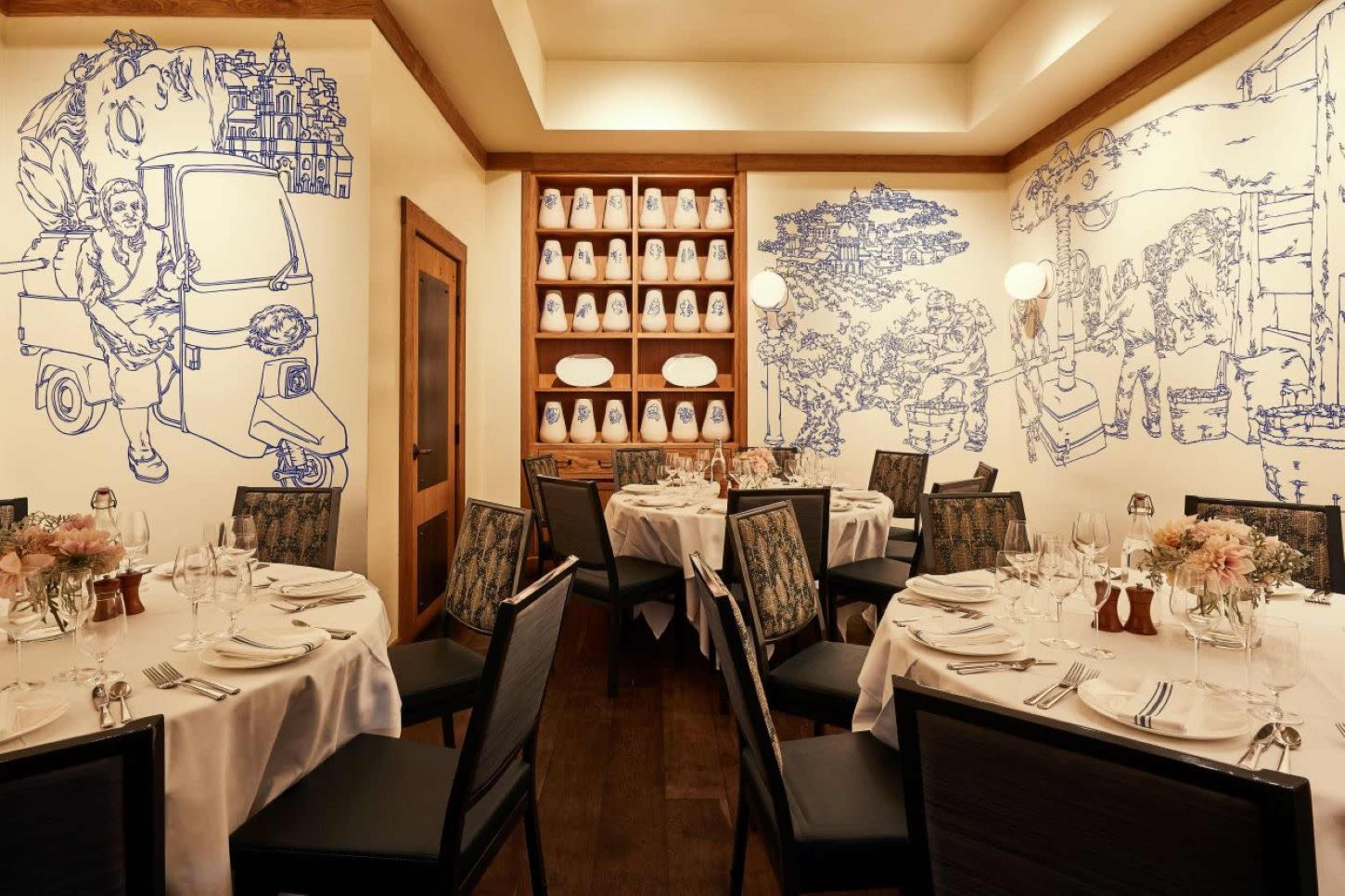 The image shows a restaurant dining area with tables set for guests and blue line art murals on the walls depicting various scenes.