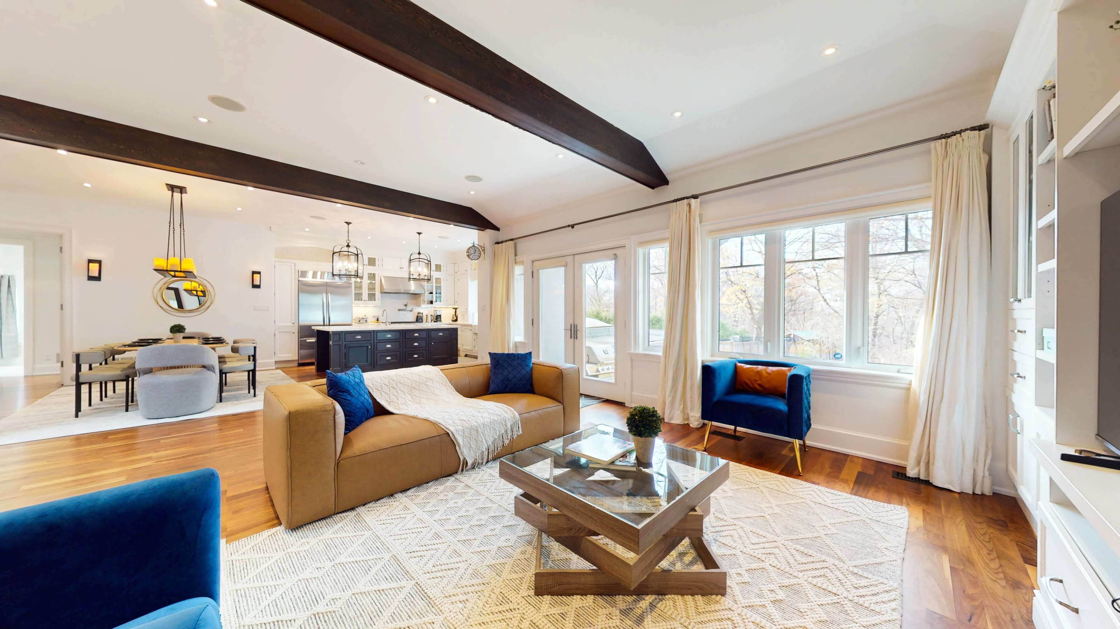 The image shows a stylish living room with a sofa, blue accent chairs, and large windows allowing natural light, connected to an open kitchen area in the background.