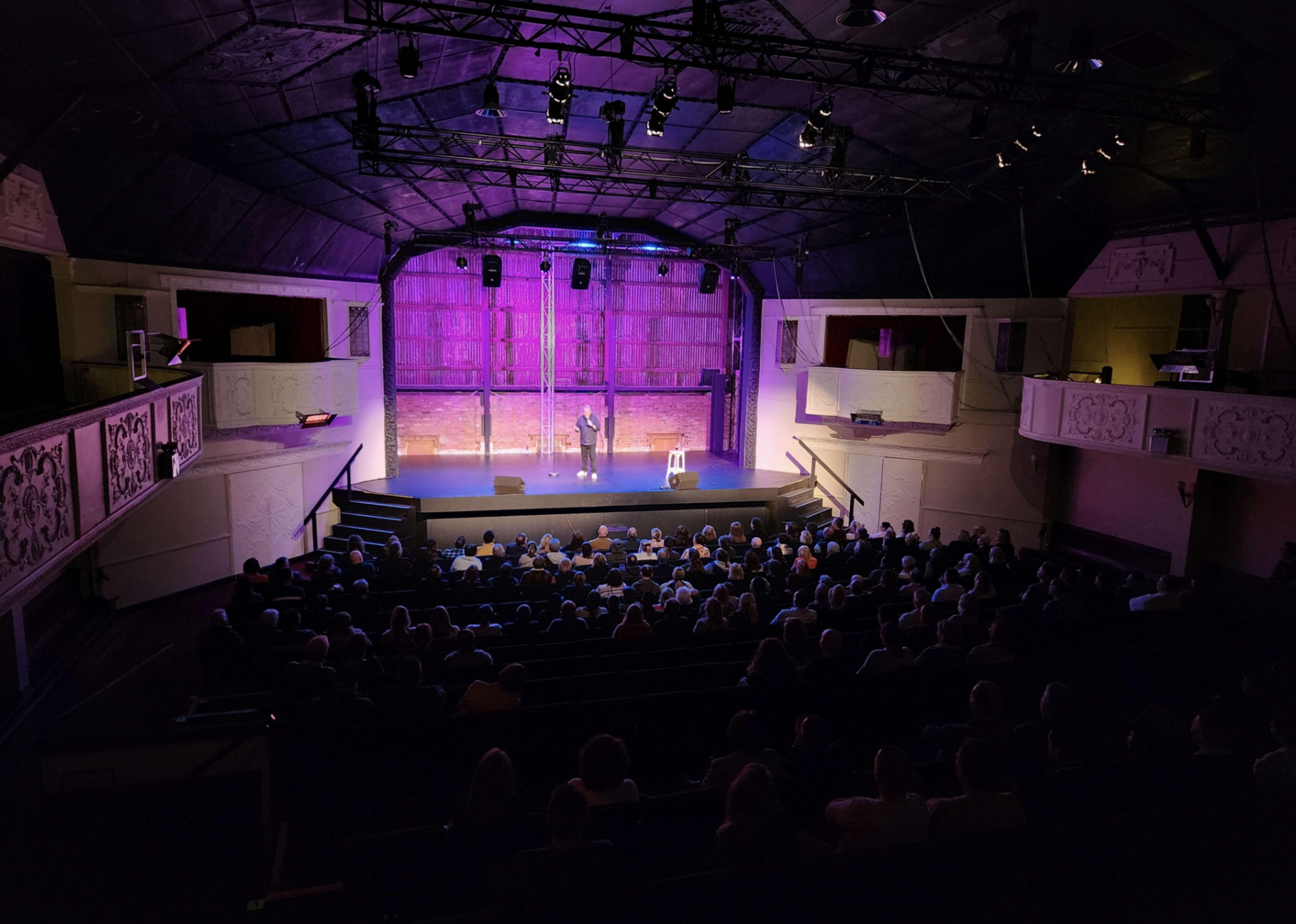 A performer stands on stage in front of a seated audience in a theater with ornate balconies and lighting.