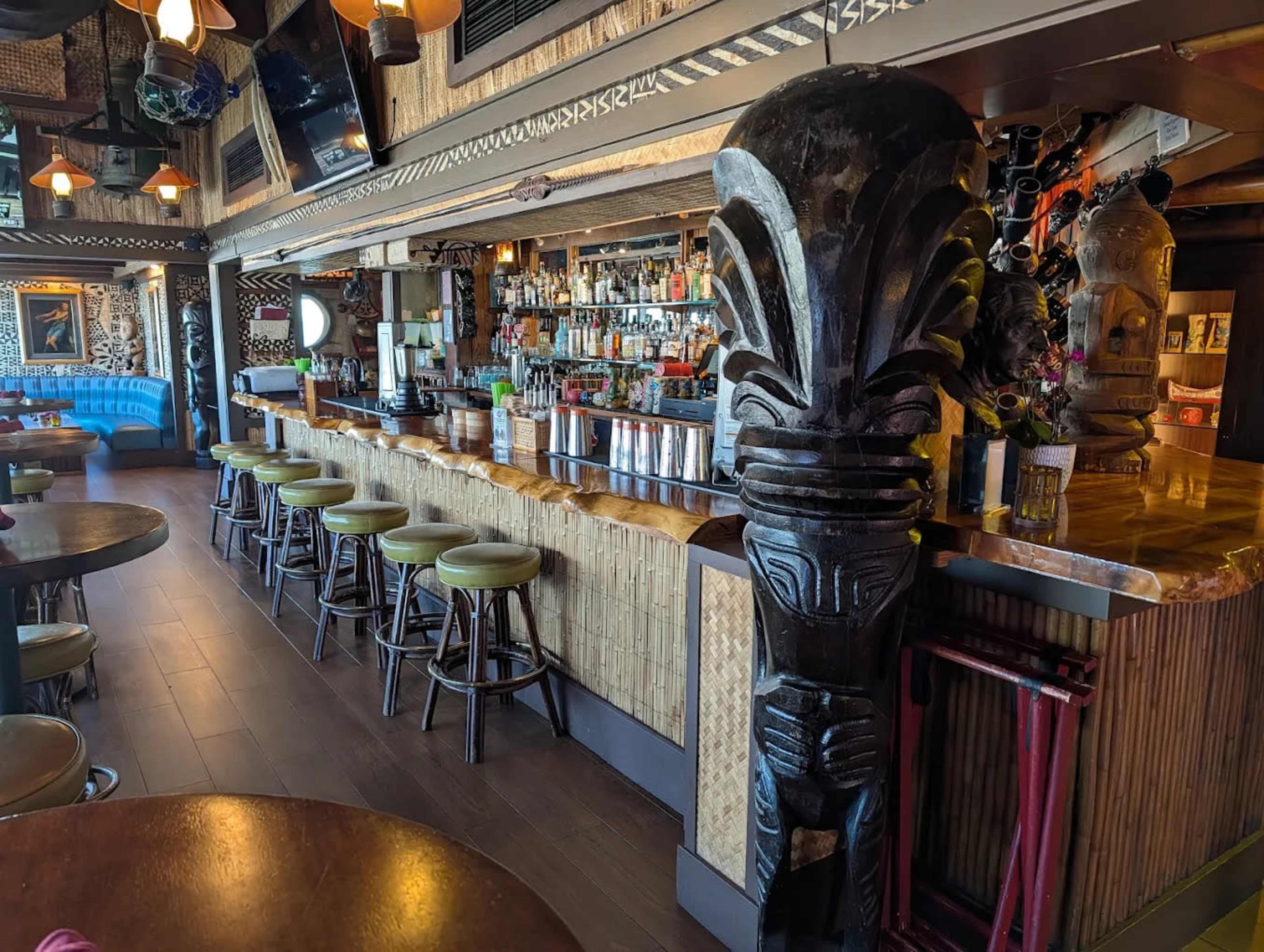 The image shows a tropical-themed bar with wooden stools, a glossy countertop, and carved tiki sculptures, surrounded by bamboo decor and shelves stocked with various bottles.