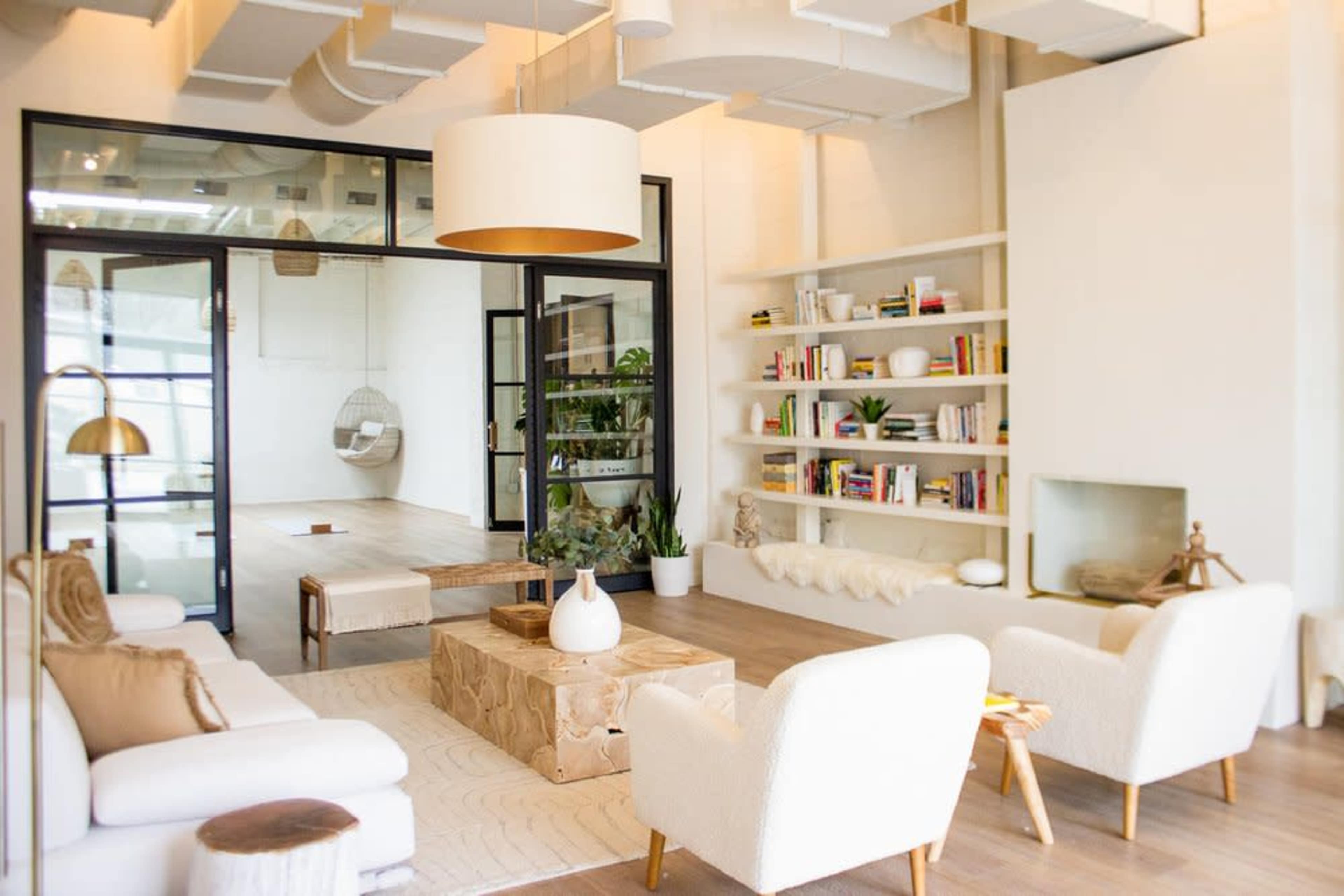 A modern living room with a light color scheme, featuring a pair of white armchairs, a wooden coffee table, and shelves filled with books and decorative items.