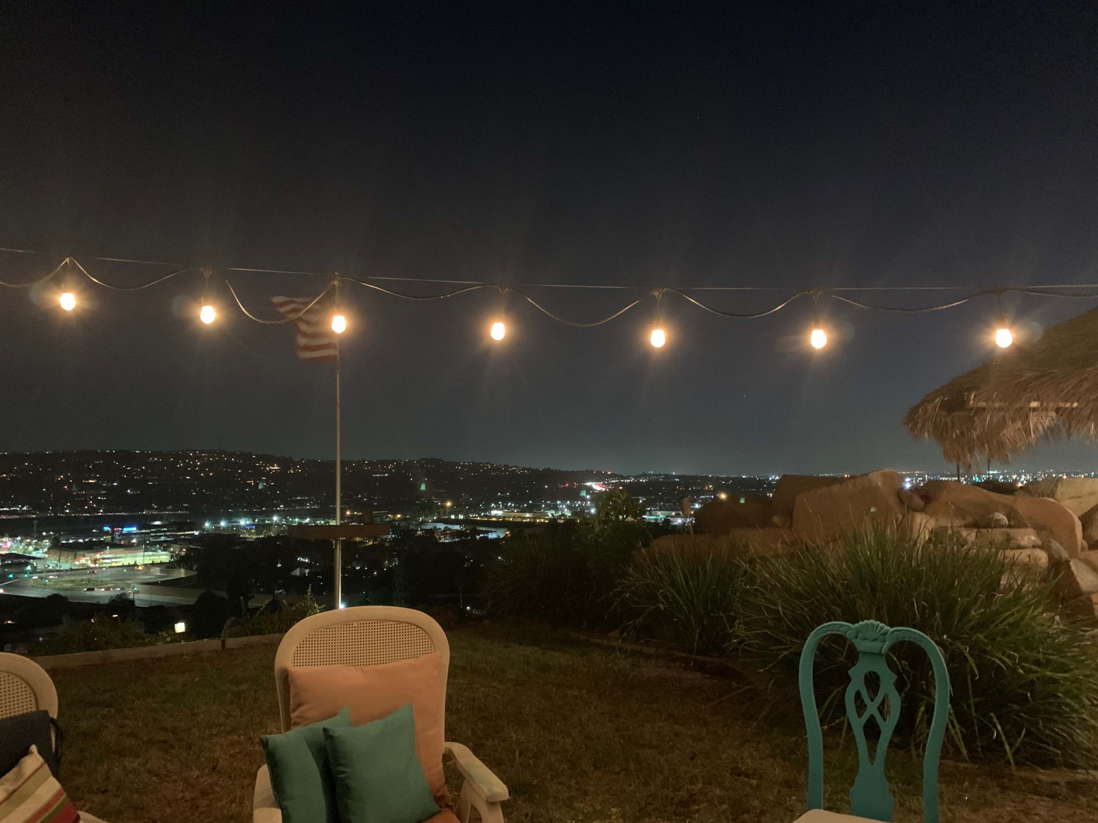 The scene features a patio with string lights overhead, overlooking a cityscape at night with a flag visible in the background.