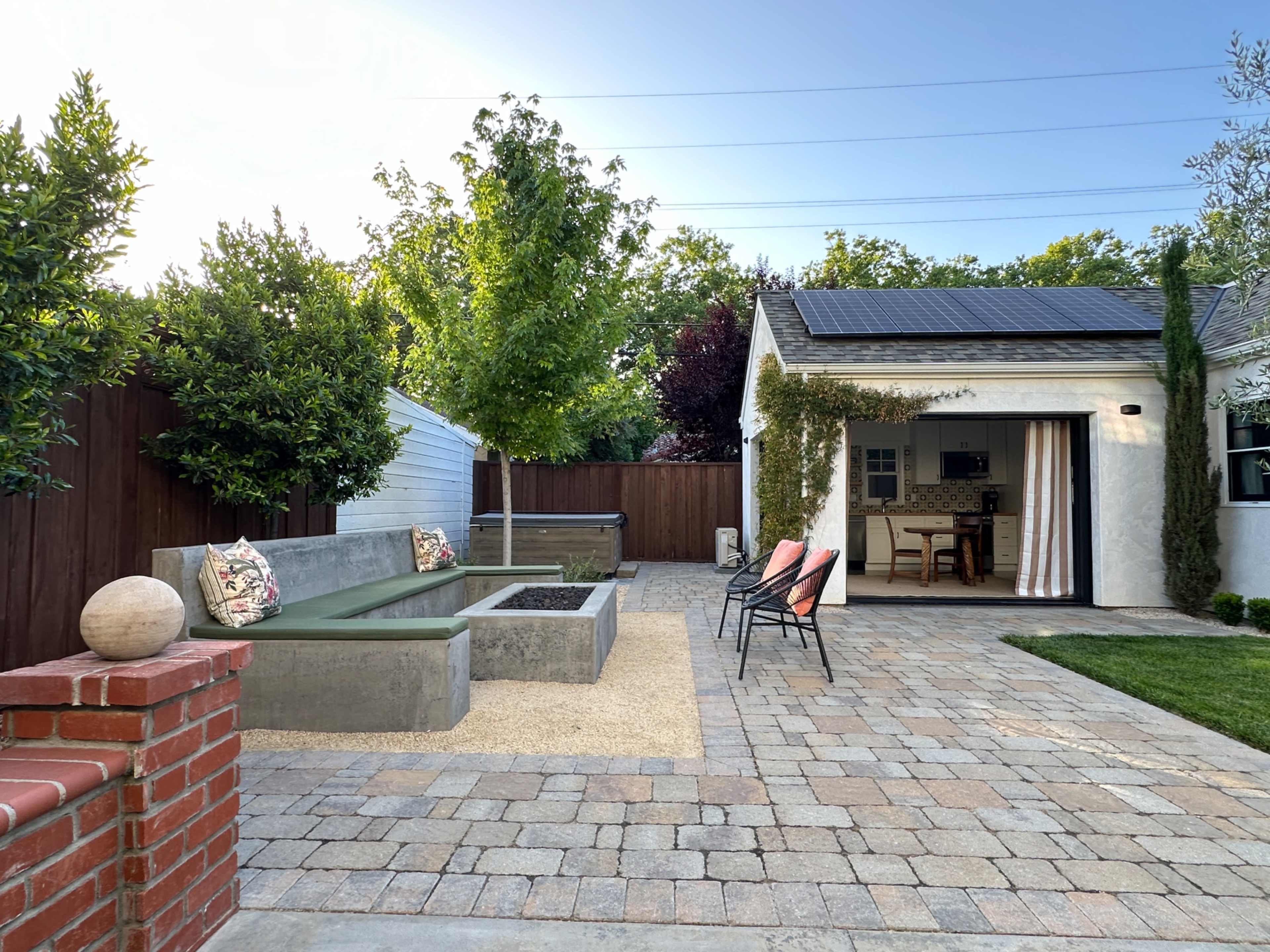 The image shows a backyard patio with a seating area made of concrete, a small garden, and a house with a light-colored exterior.