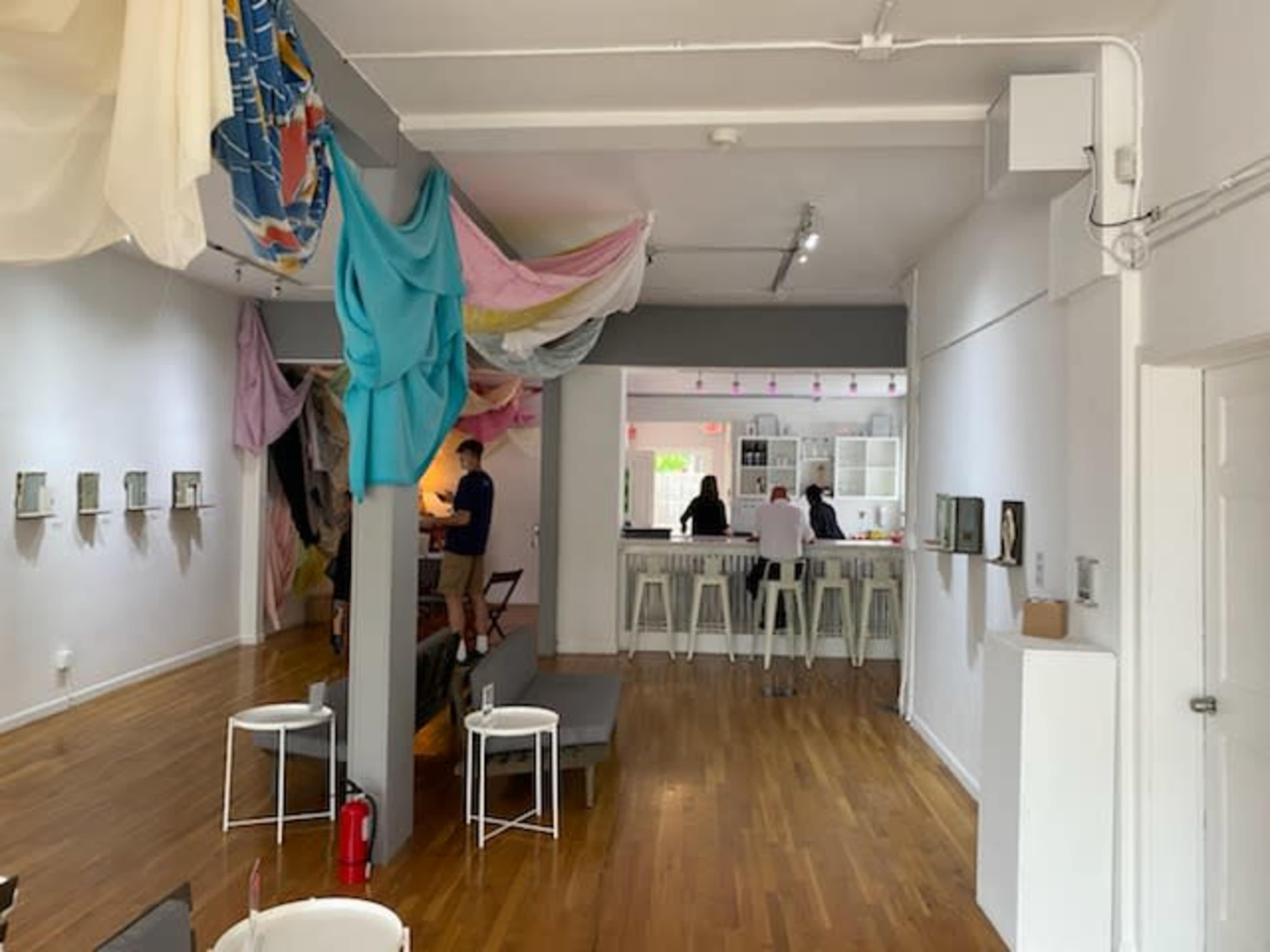 A spacious gallery or café with colorful draped fabrics overhead, a counter in the background, and several people engaged in activities.