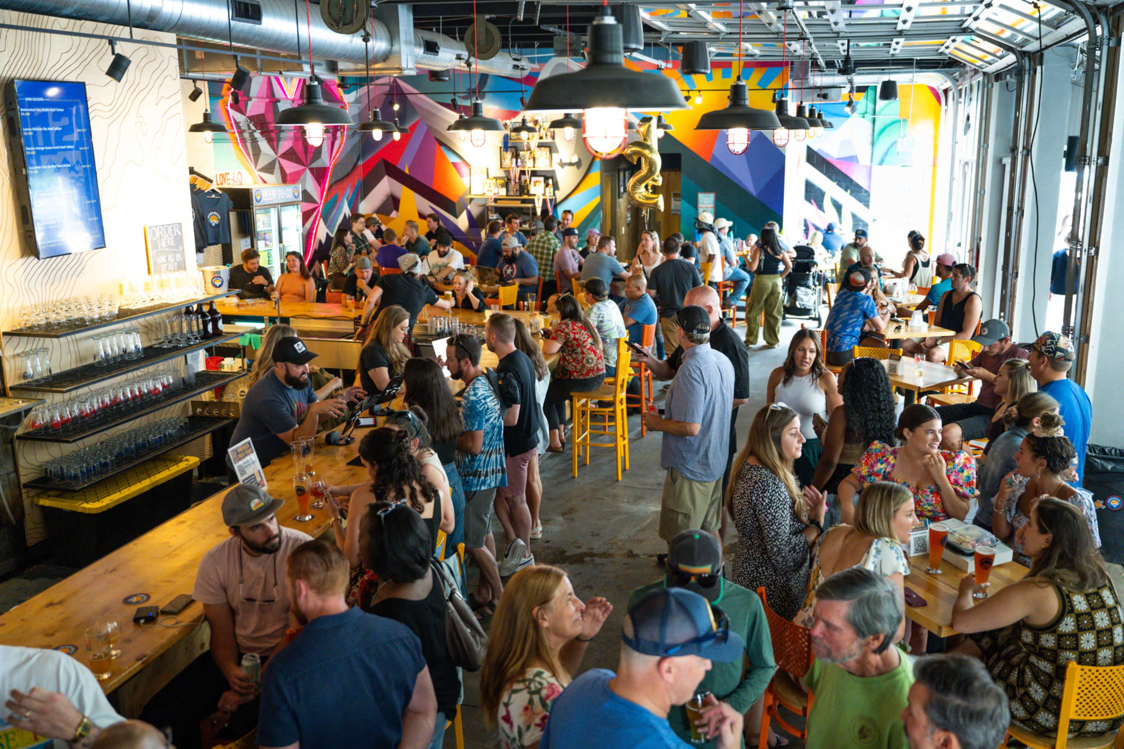 The image shows a crowded bar with patrons socializing, colorful murals on the walls, and a variety of seating arrangements.