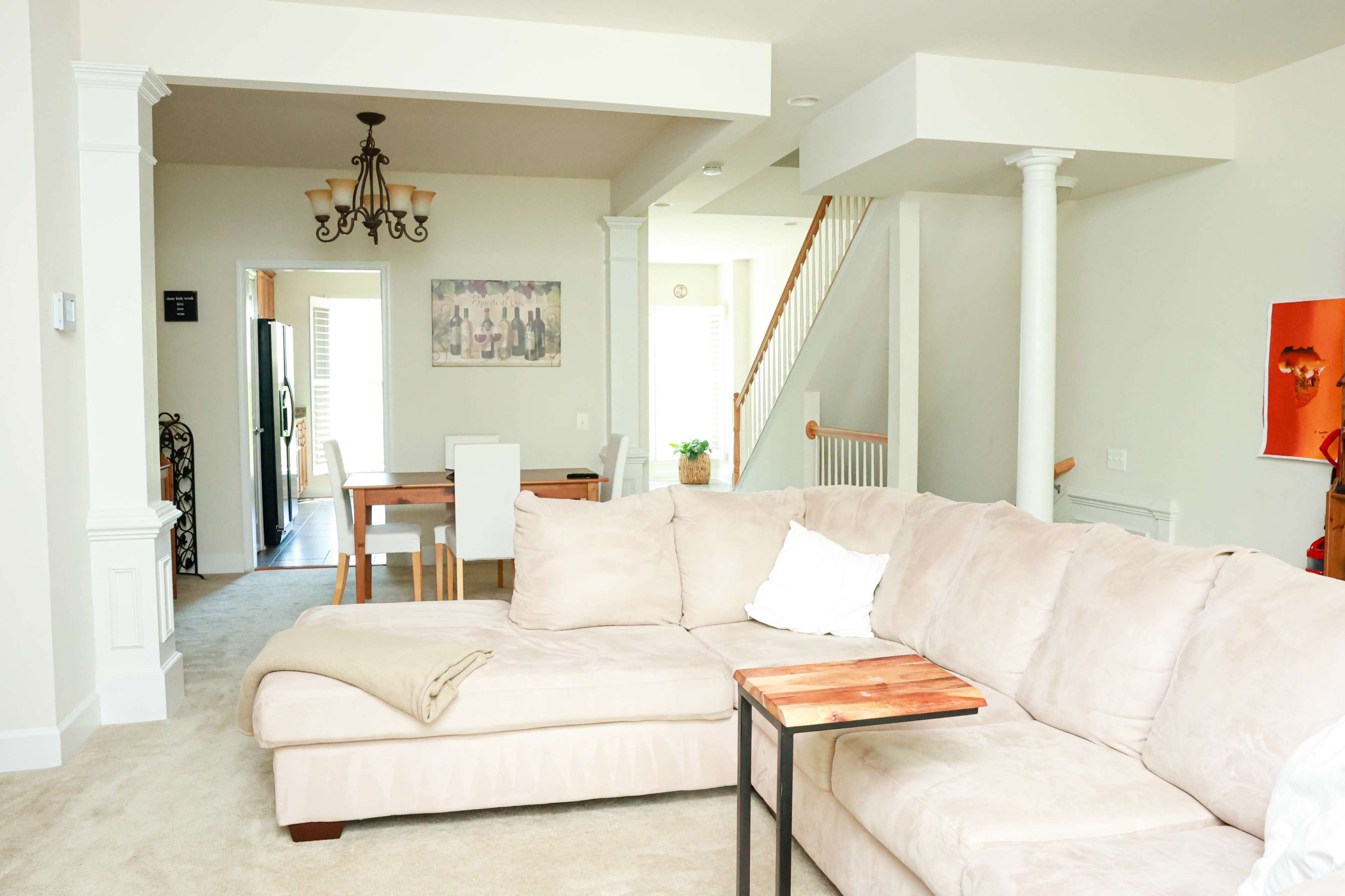 The image shows a light-filled living area featuring a beige sectional sofa, a small wooden side table, and a dining area in the background with a staircase leading to the upper level.