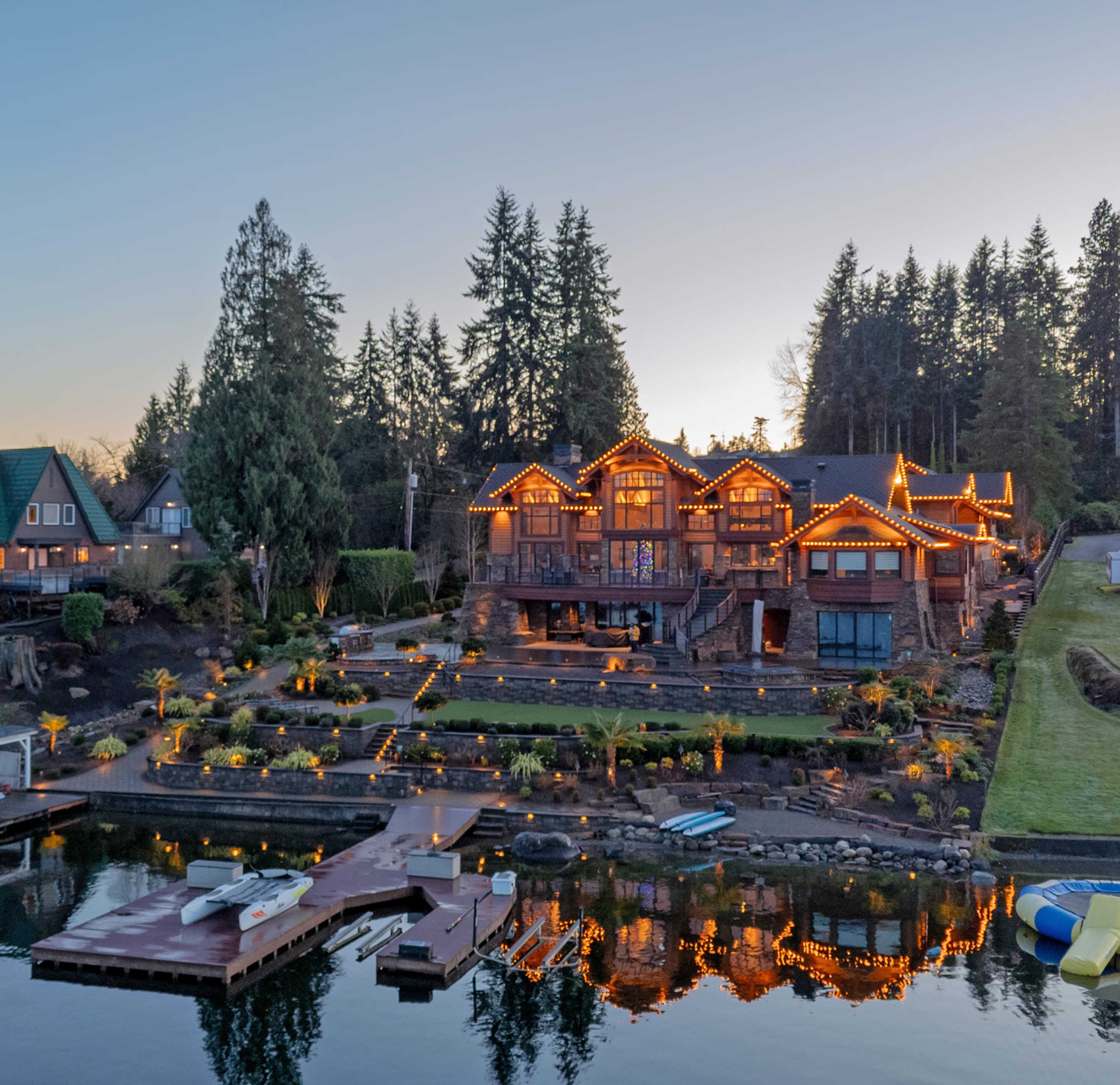 A large, illuminated waterfront house is surrounded by landscaped gardens and a dock, with trees and hills in the background.