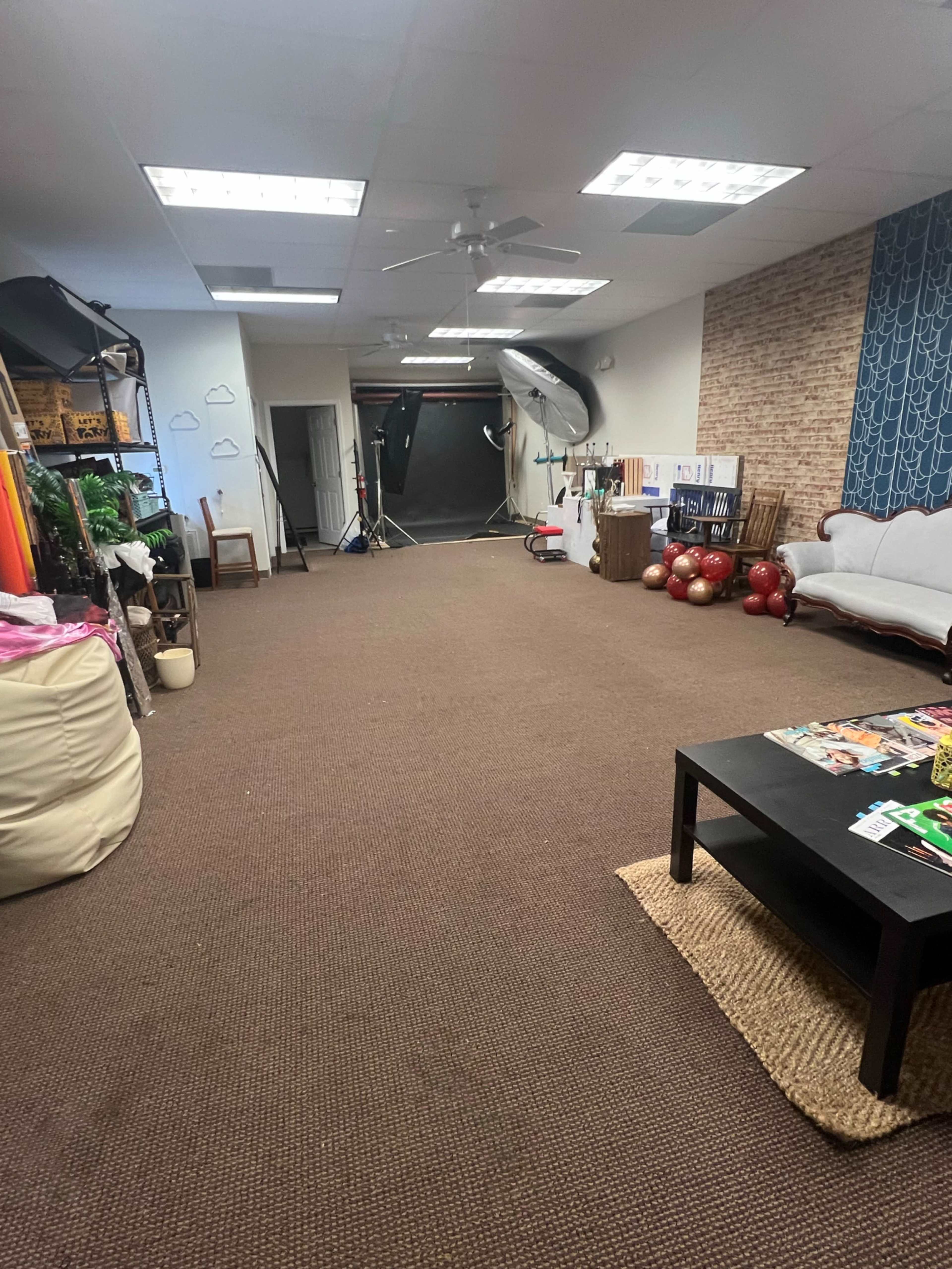 The image shows a spacious room with a beige carpet, featuring a brown sofa, a coffee table, various props on the sides, and photography equipment in the background.
