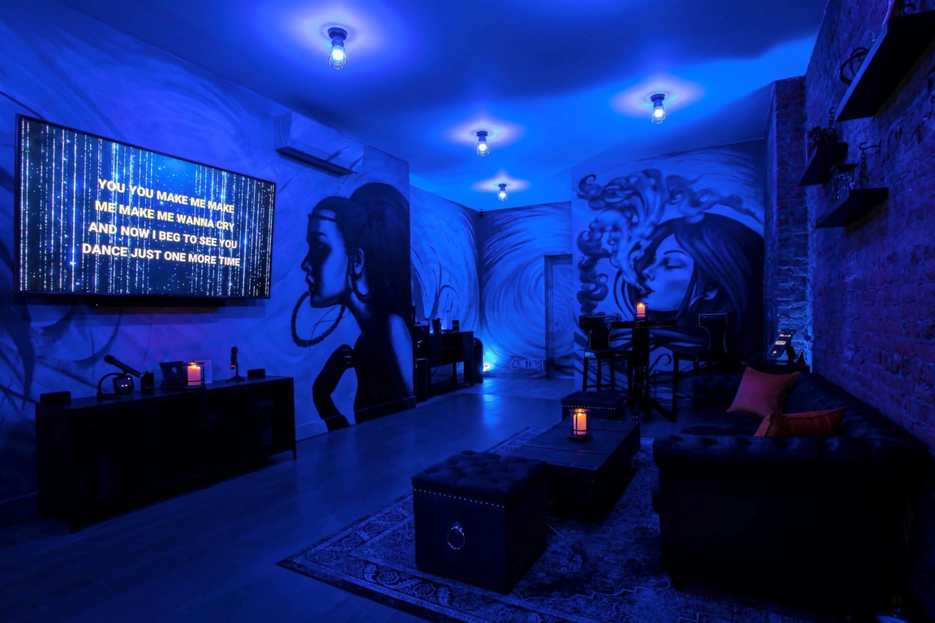 The room features blue lighting, a large television displaying lyrics, and walls adorned with artistic murals of women.