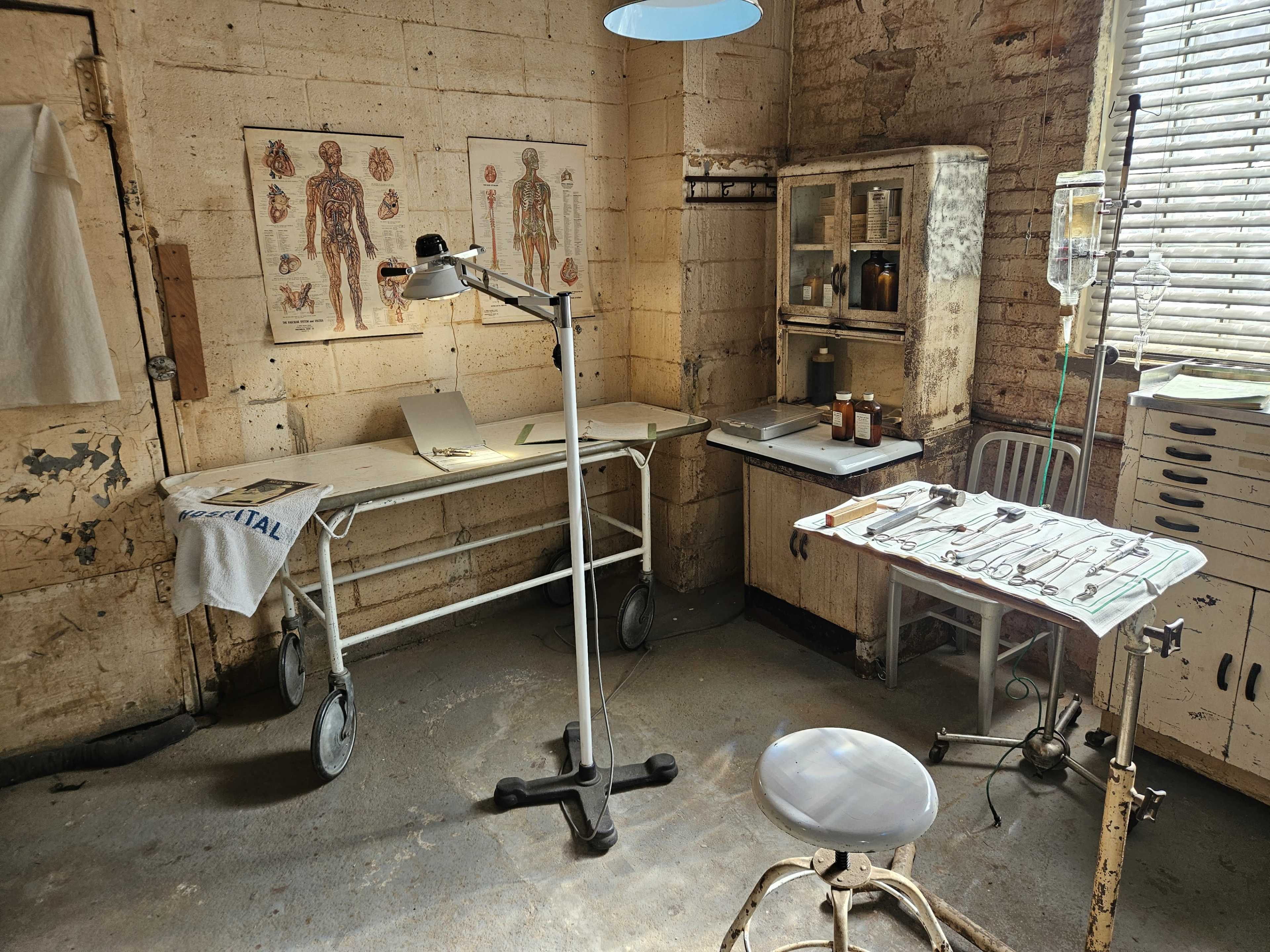 The image shows an old medical examination room with a treatment table, a desk containing various medical supplies, and anatomical posters on the wall.