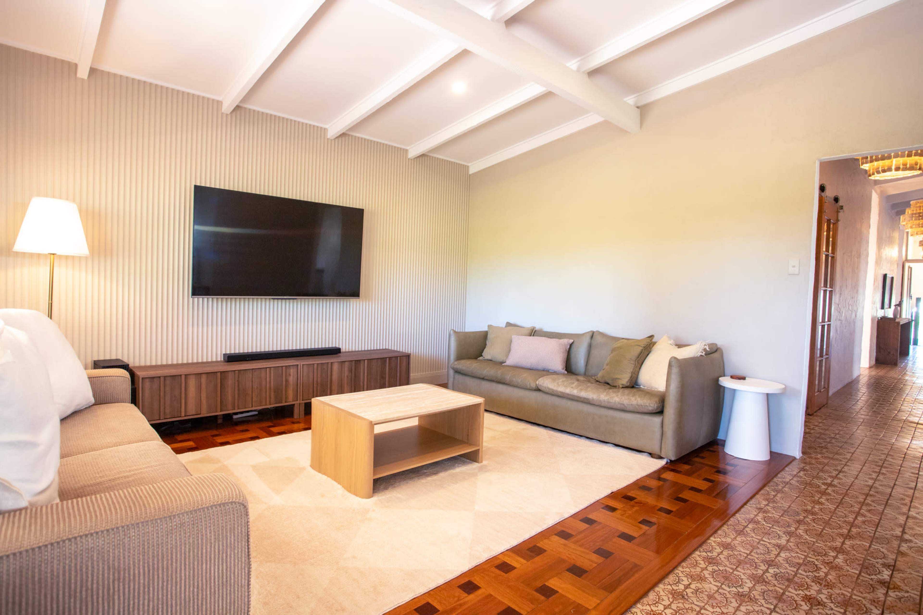 The image shows a living room with two sofas, a coffee table, a flat-screen television mounted on the wall, and a doorway leading to another space.