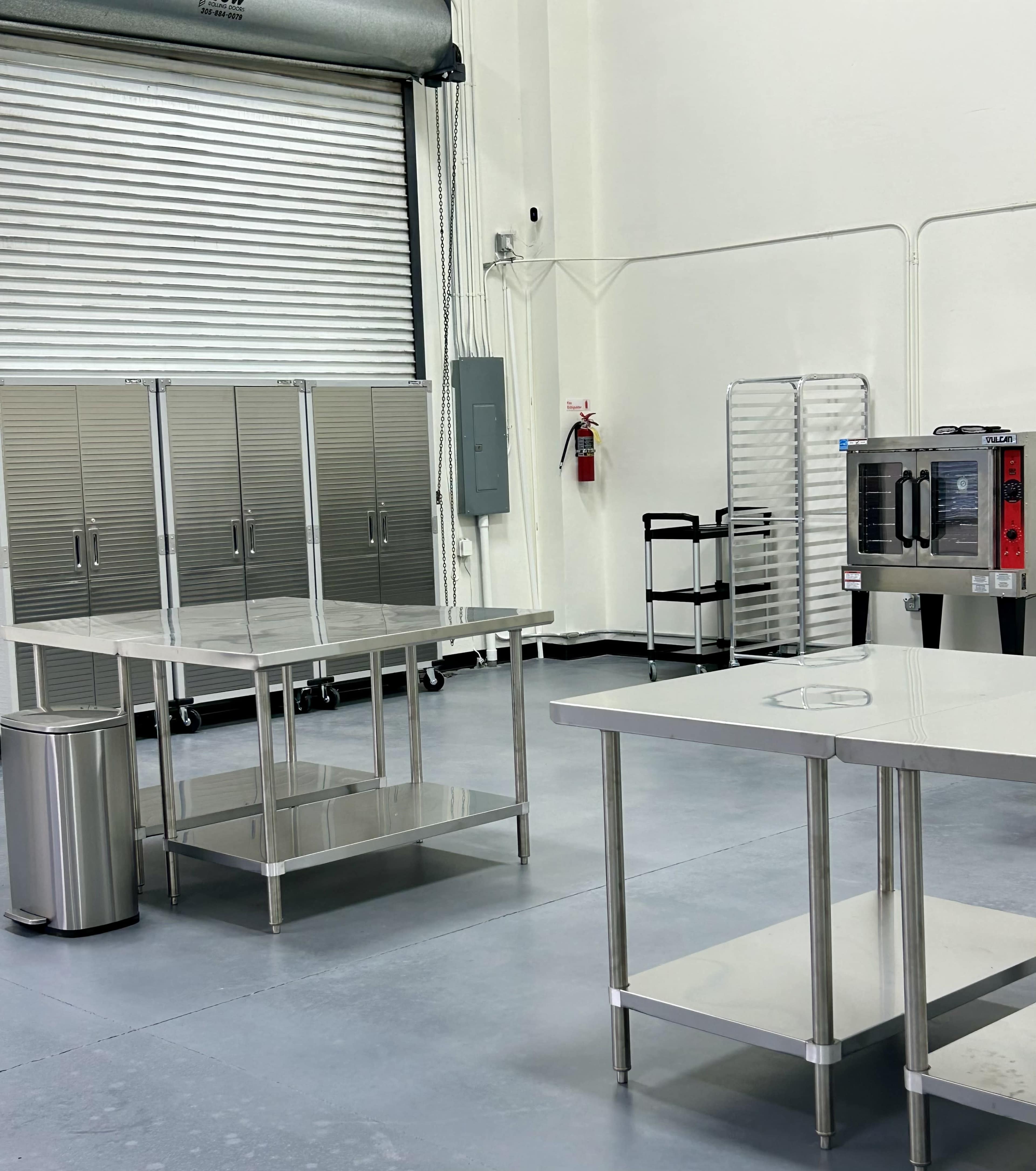 The image shows a clean, industrial kitchen space with stainless steel tables, a microwave, and storage cabinets.
