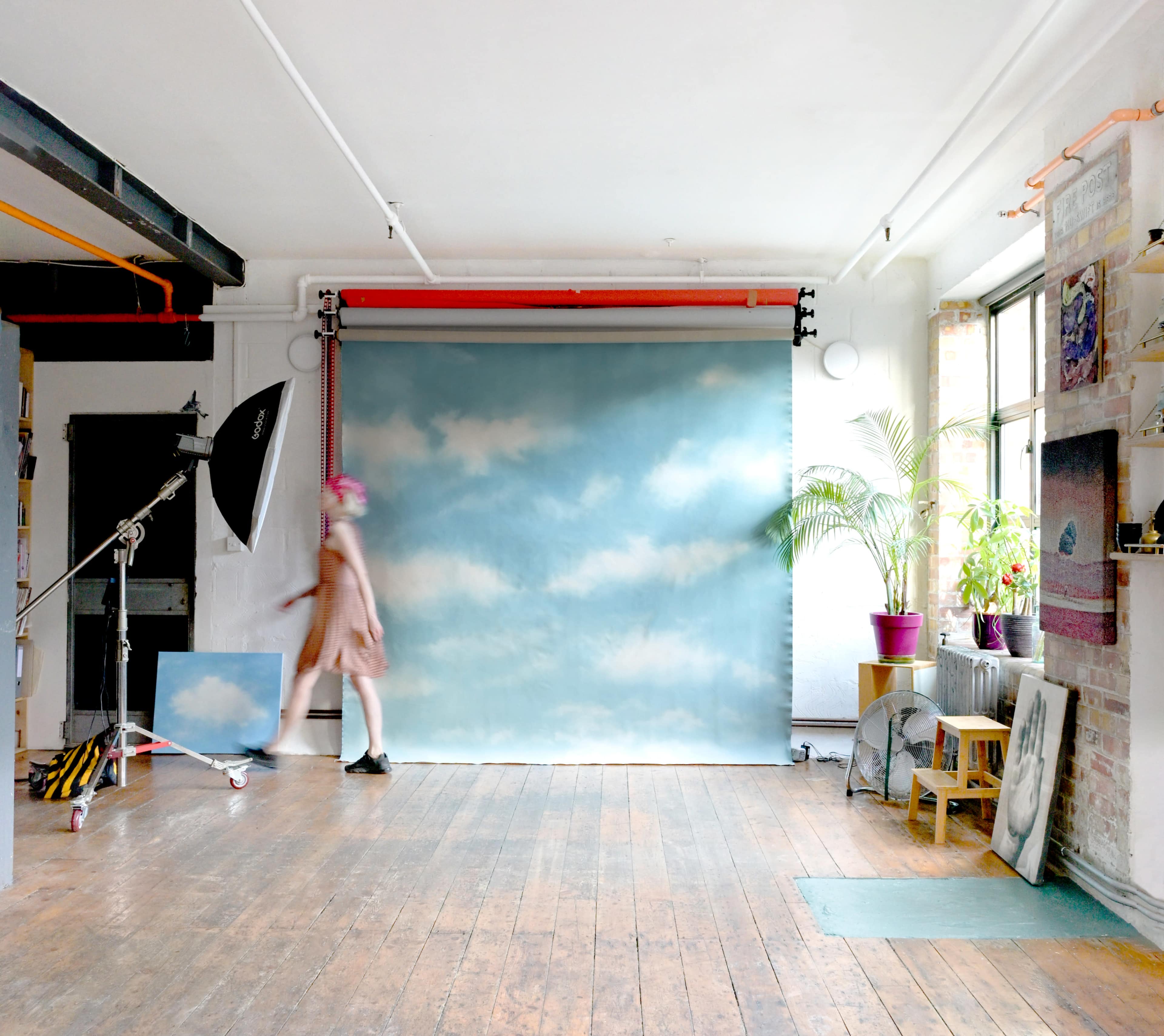 A person with pink hair walks across a studio space featuring a blue backdrop with clouds and various art supplies arranged around the room.