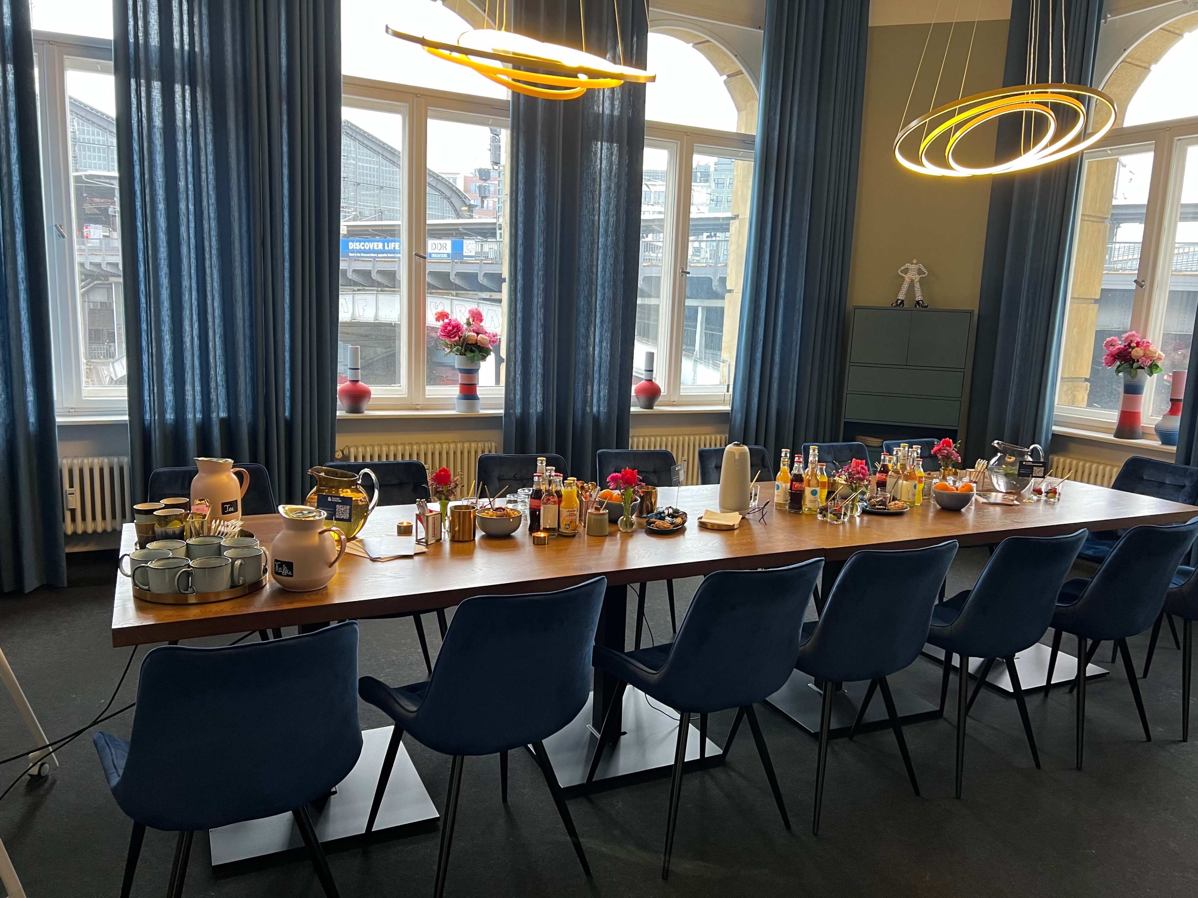 A long dining table is set with various drinks and snacks in a room with large windows and blue curtains.
