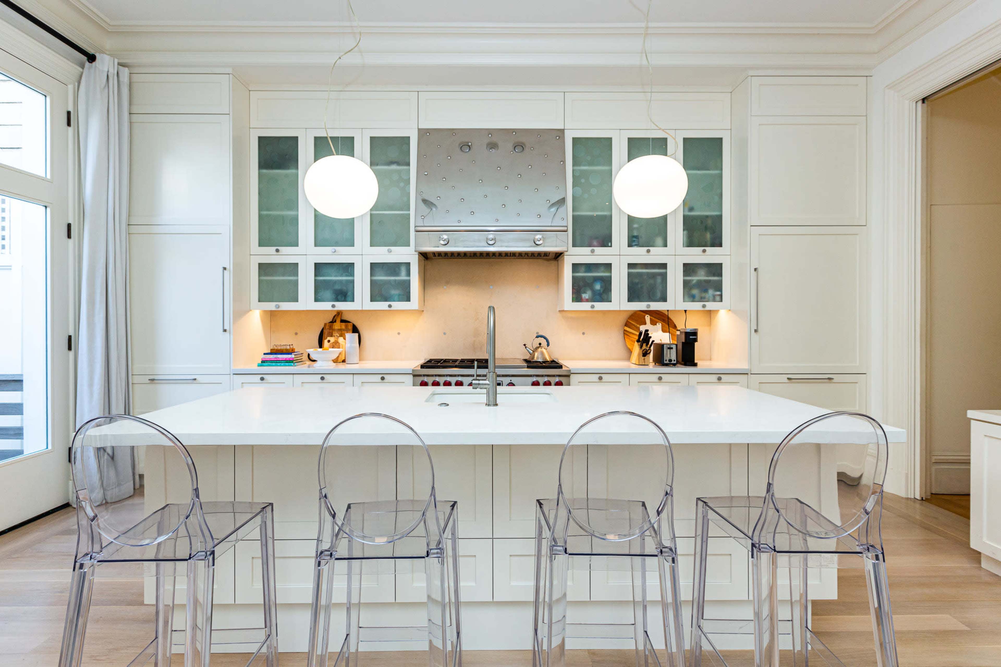 A modern kitchen features a large white island with four clear acrylic bar stools and a stainless steel range hood above a cooking area.