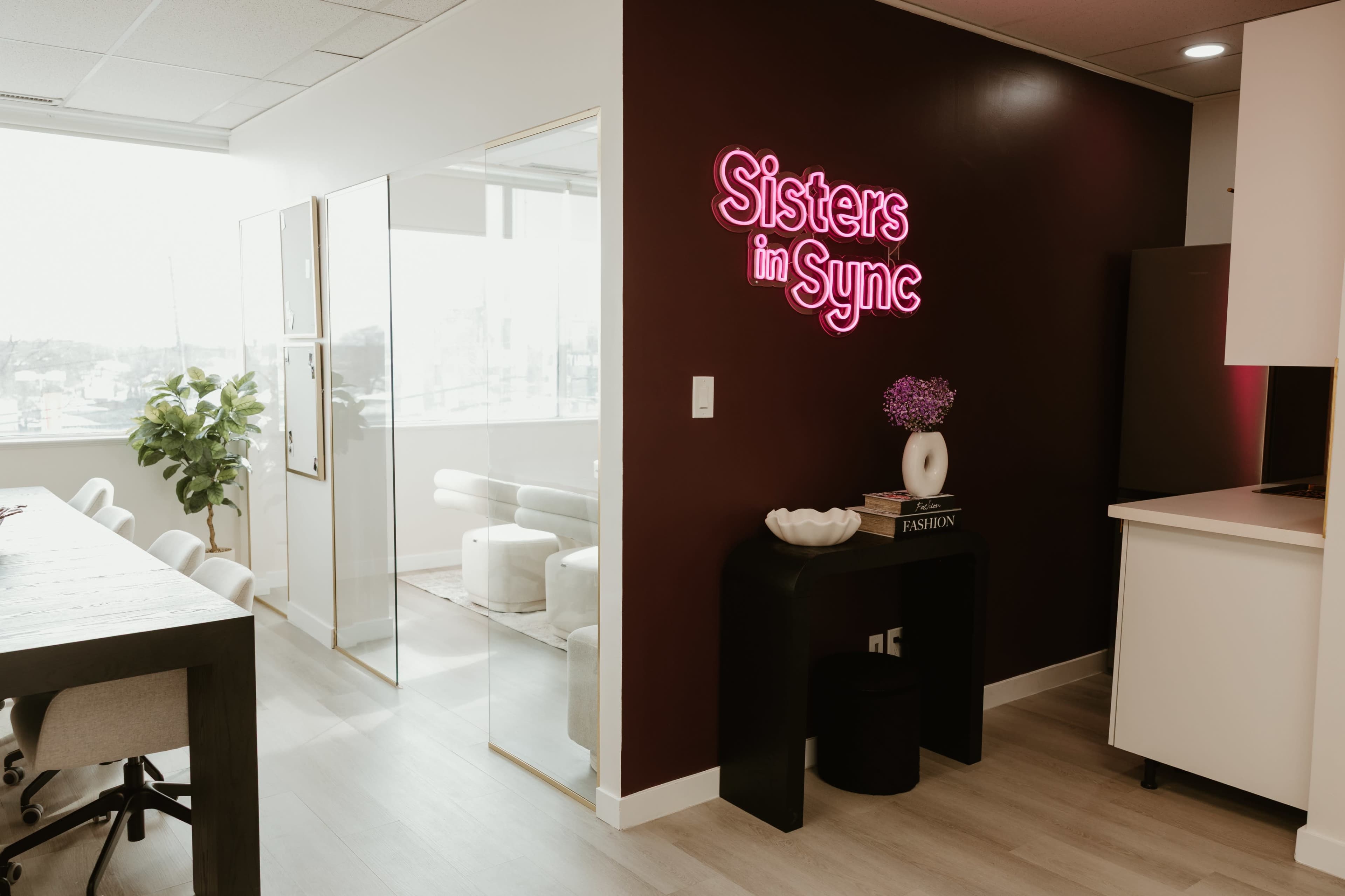 The image shows a modern office space with a glowing neon sign that reads "Sisters in Sync," a glass-walled room, and a light-colored wooden table with chairs.