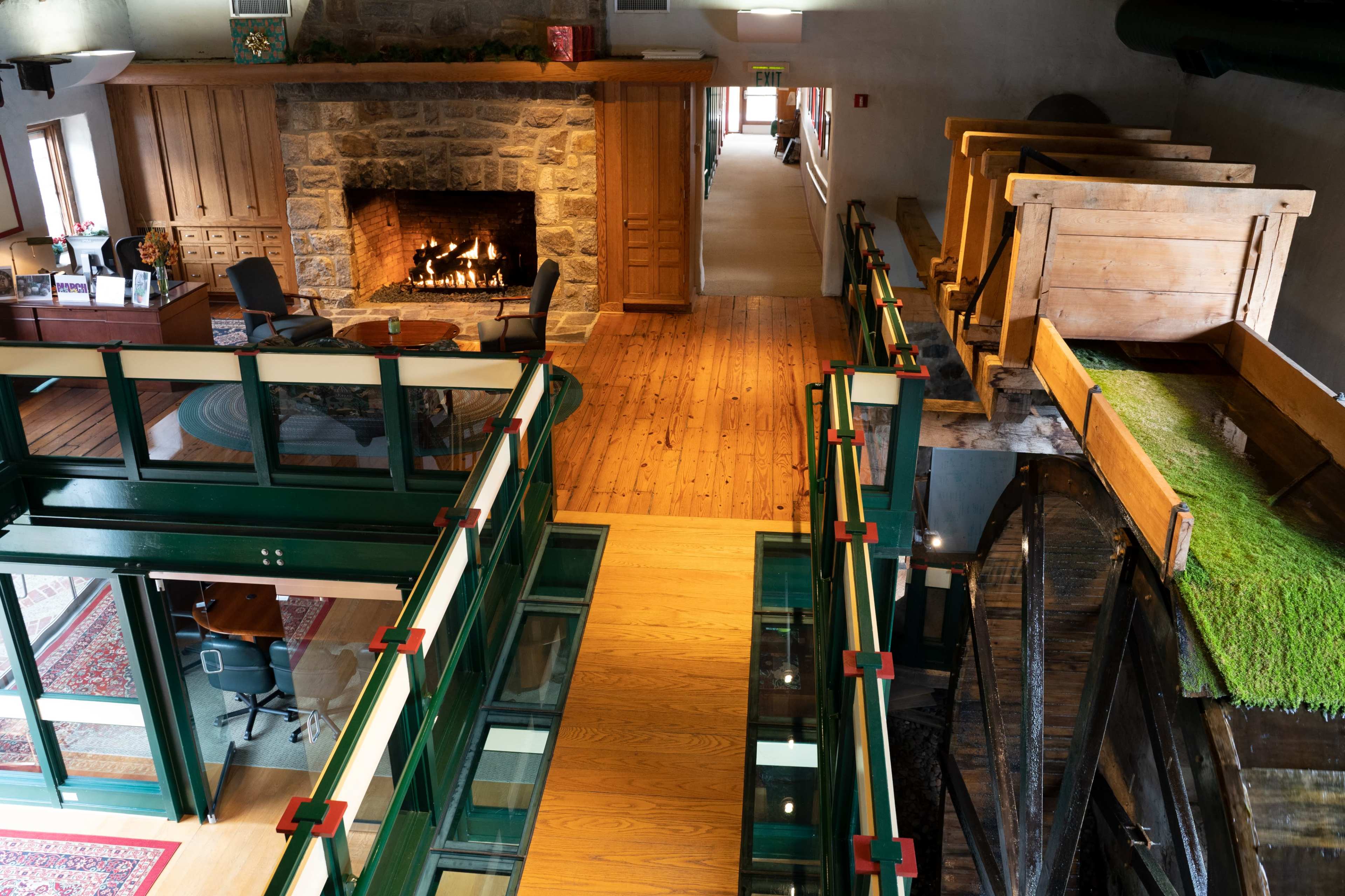 An interior space featuring a large stone fireplace, wooden floors, and a glass walkway overlooking a structure with wooden boxes and greenery.