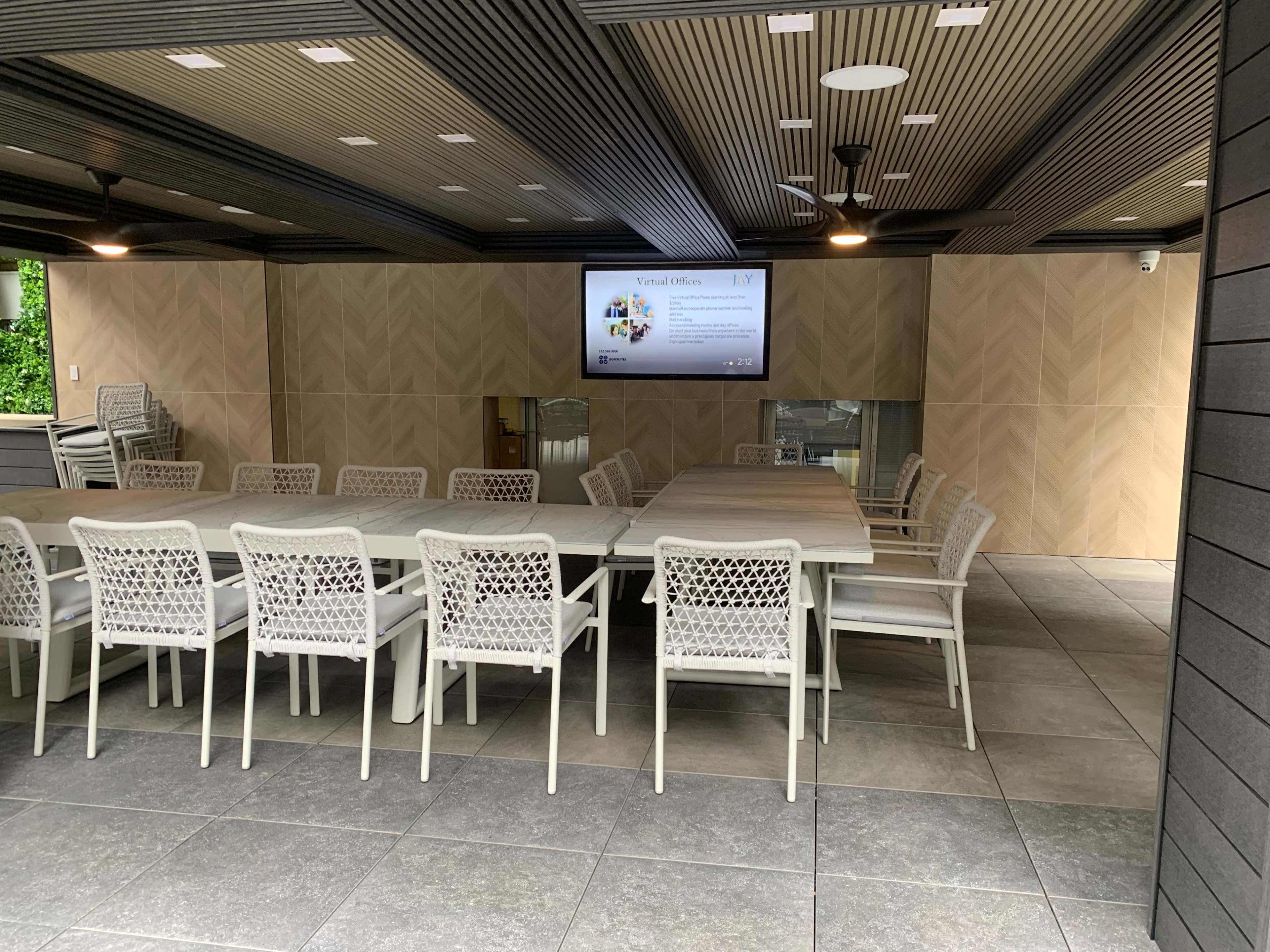 The image shows a modern, covered outdoor meeting space with a large table surrounded by white chairs and a screen on the wall.