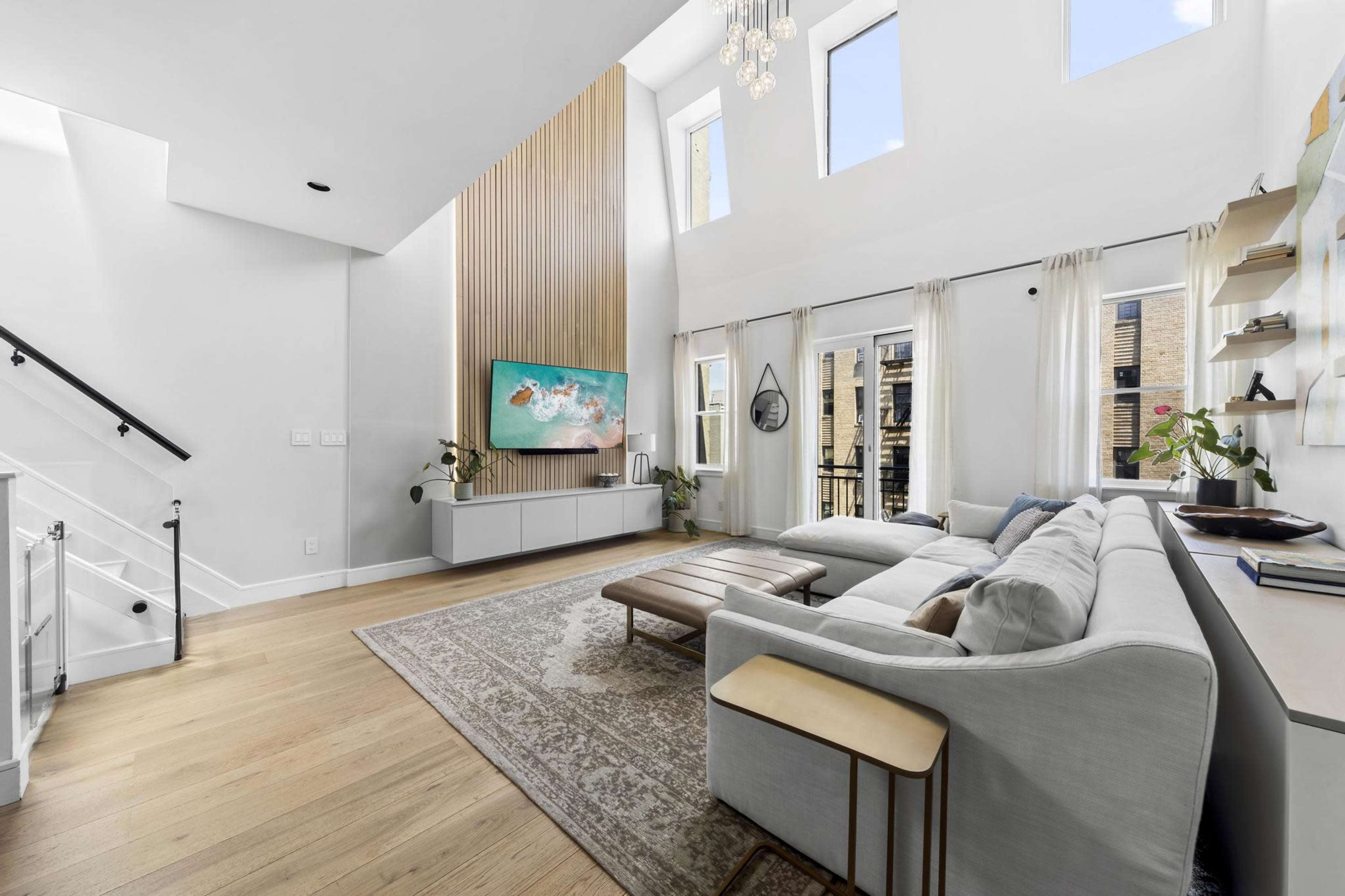 A modern living room with a high ceiling, large windows, a sectional sofa, and a wall-mounted TV above a white media console.