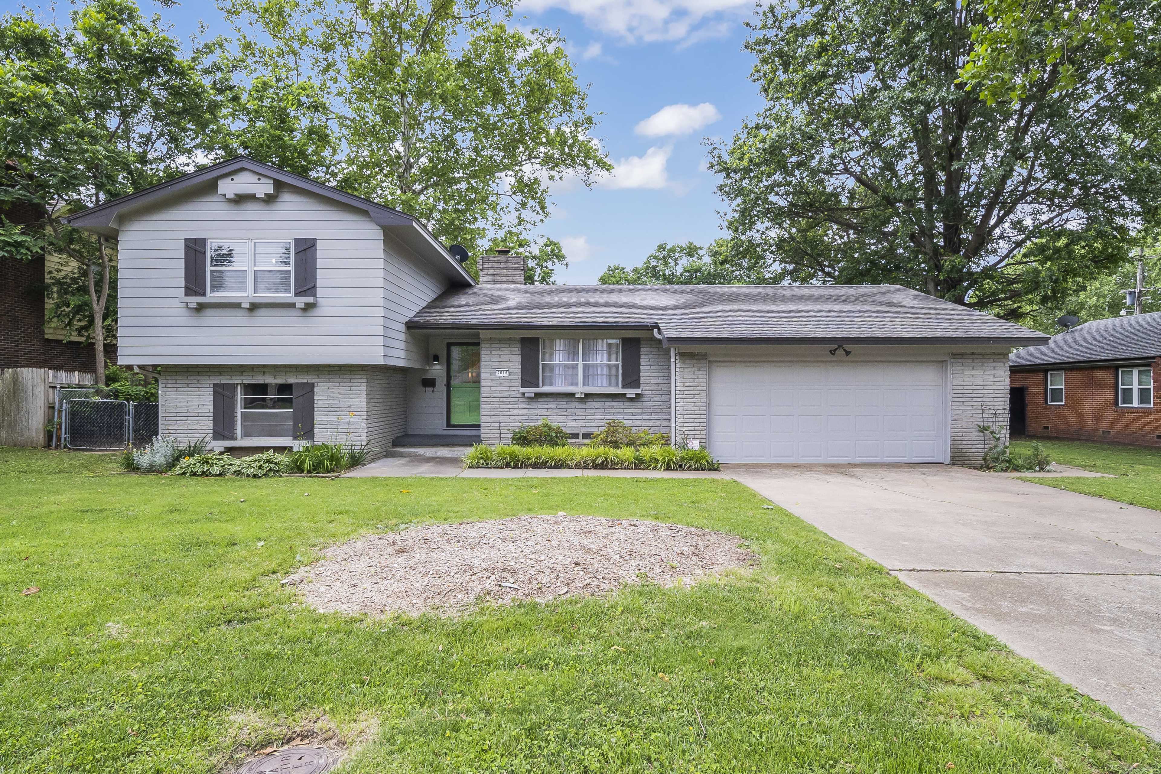 A two-story gray house with a green front door and a large front yard, featuring a circular stone garden and a concrete driveway.