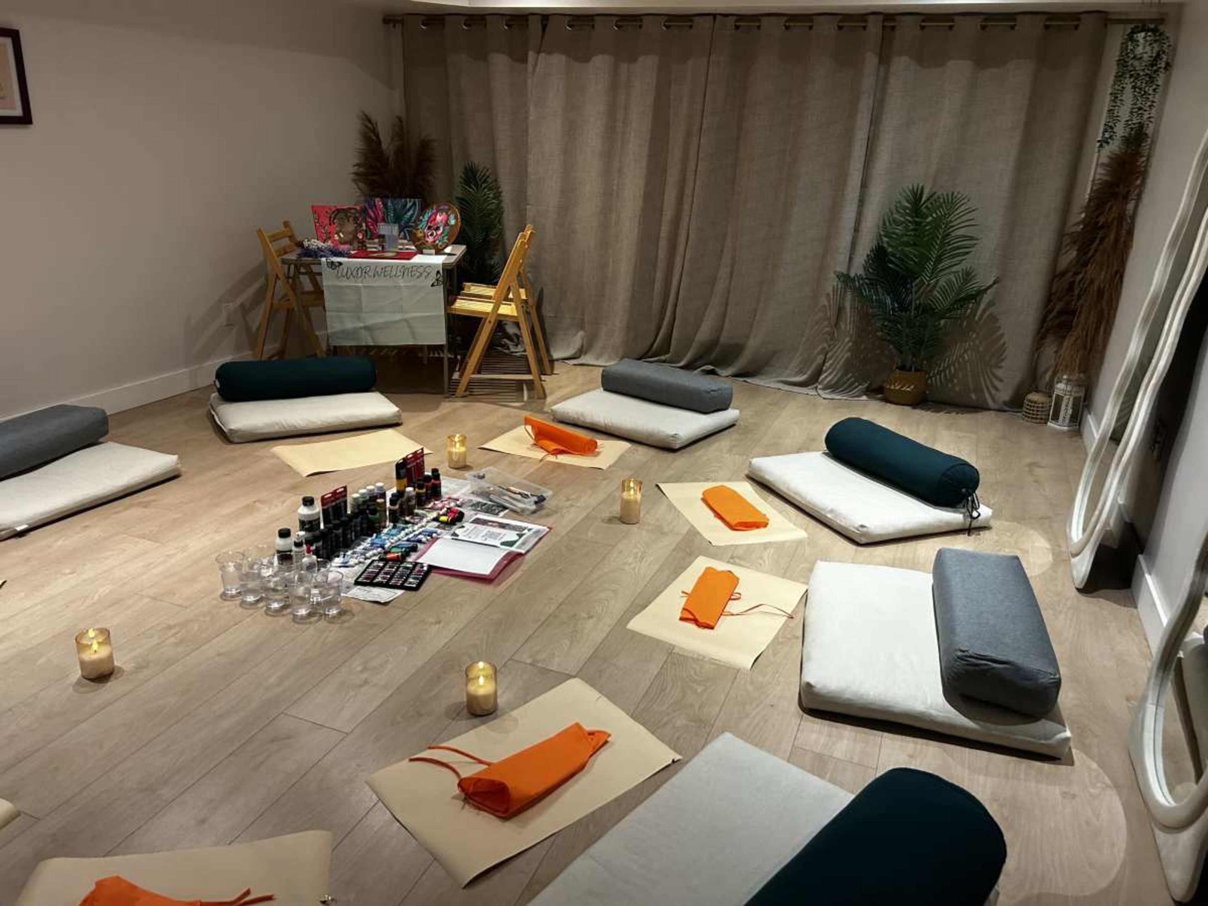 The image shows a meditation or wellness room with floor cushions arranged in a circle, candles lit, and a table set up at the back with art supplies and decorative items.