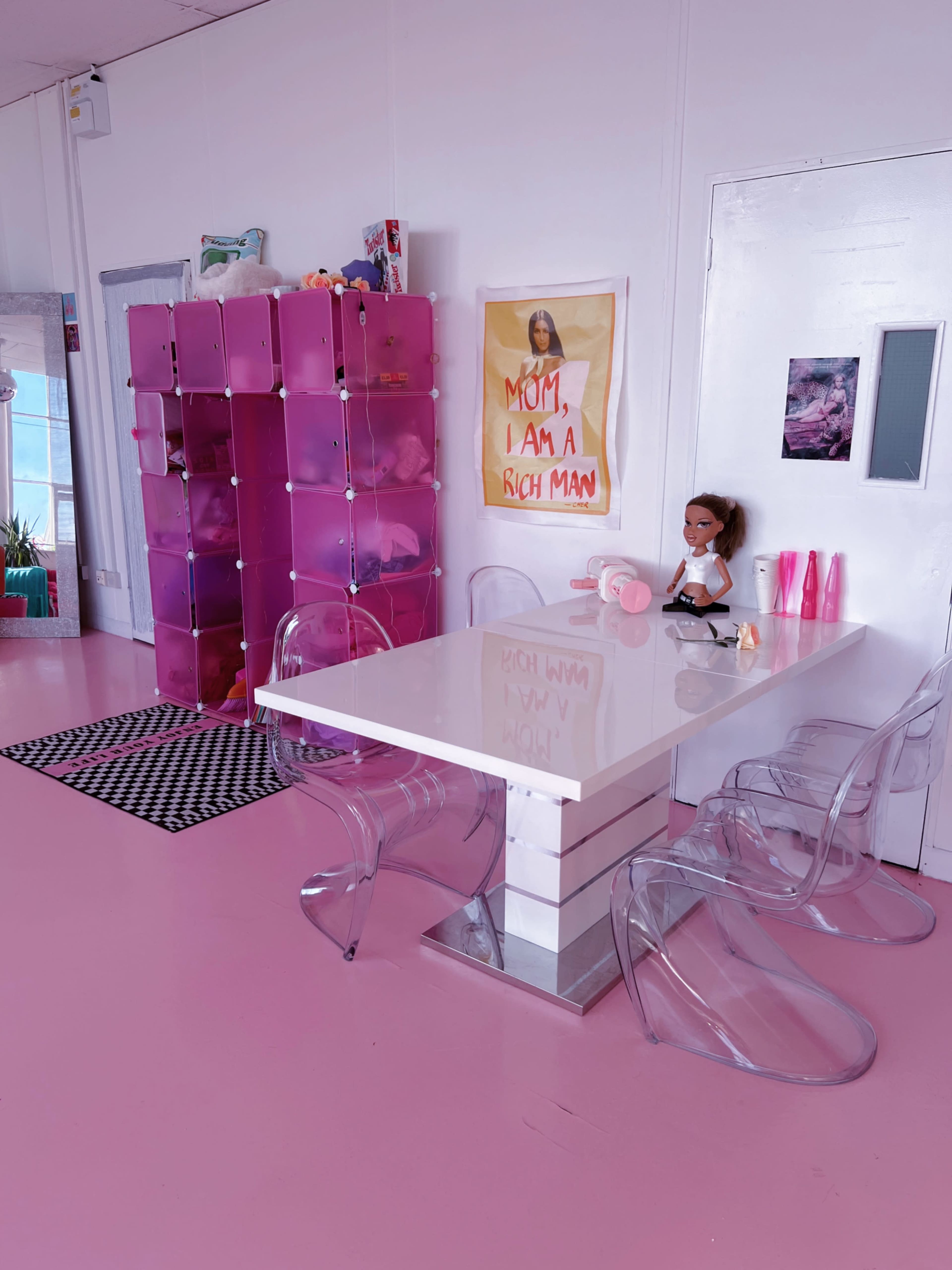 A bright pink room features a white table surrounded by transparent chairs, with a pink storage unit and a poster on the wall.