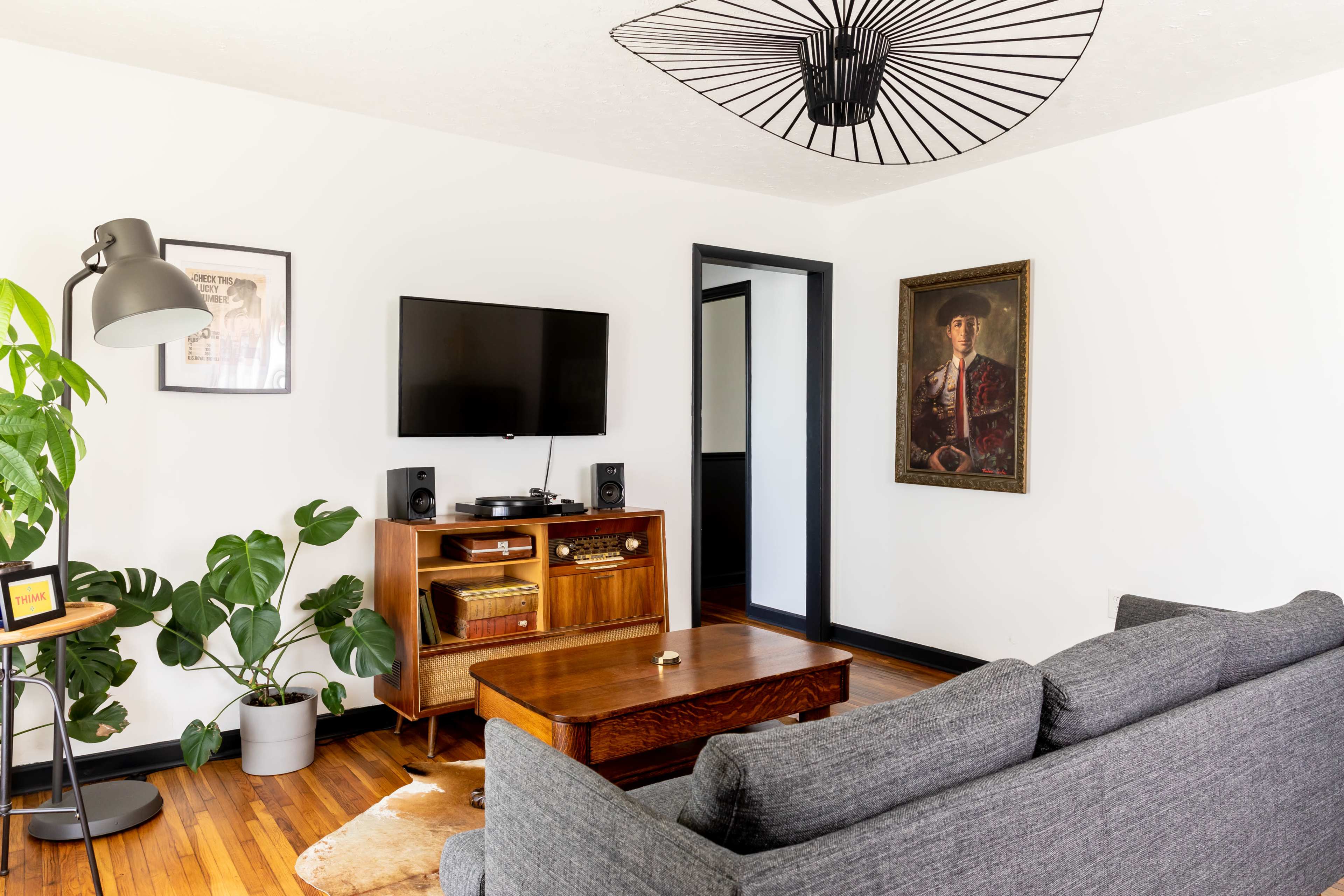 The living room features a gray sofa, a wooden coffee table, a wall-mounted television, a vintage portrait, and a large potted plant near a modern floor lamp.