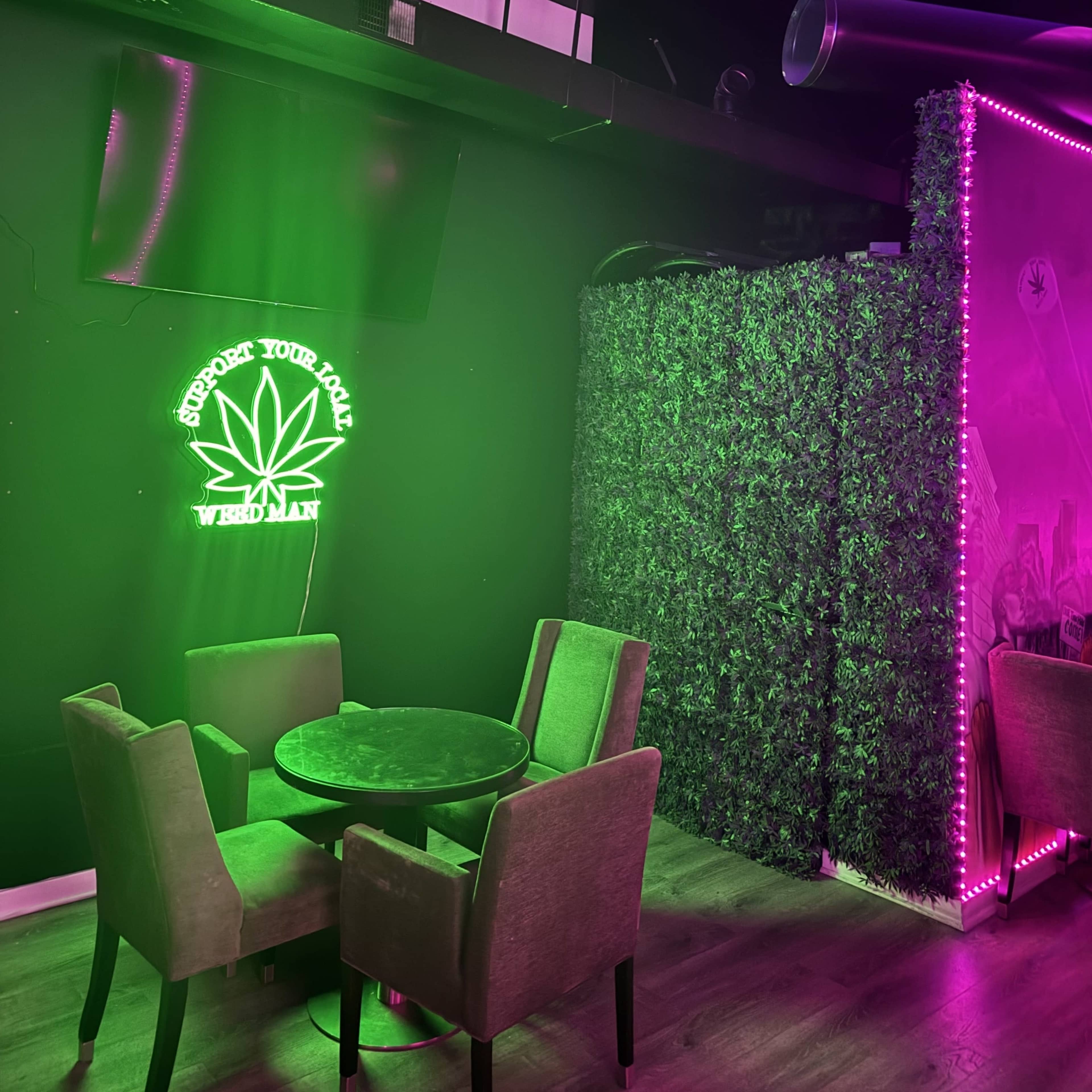 The image shows a seating area with four chairs around a round table, a neon sign featuring a cannabis leaf on the wall, and a green wall covered in faux foliage.