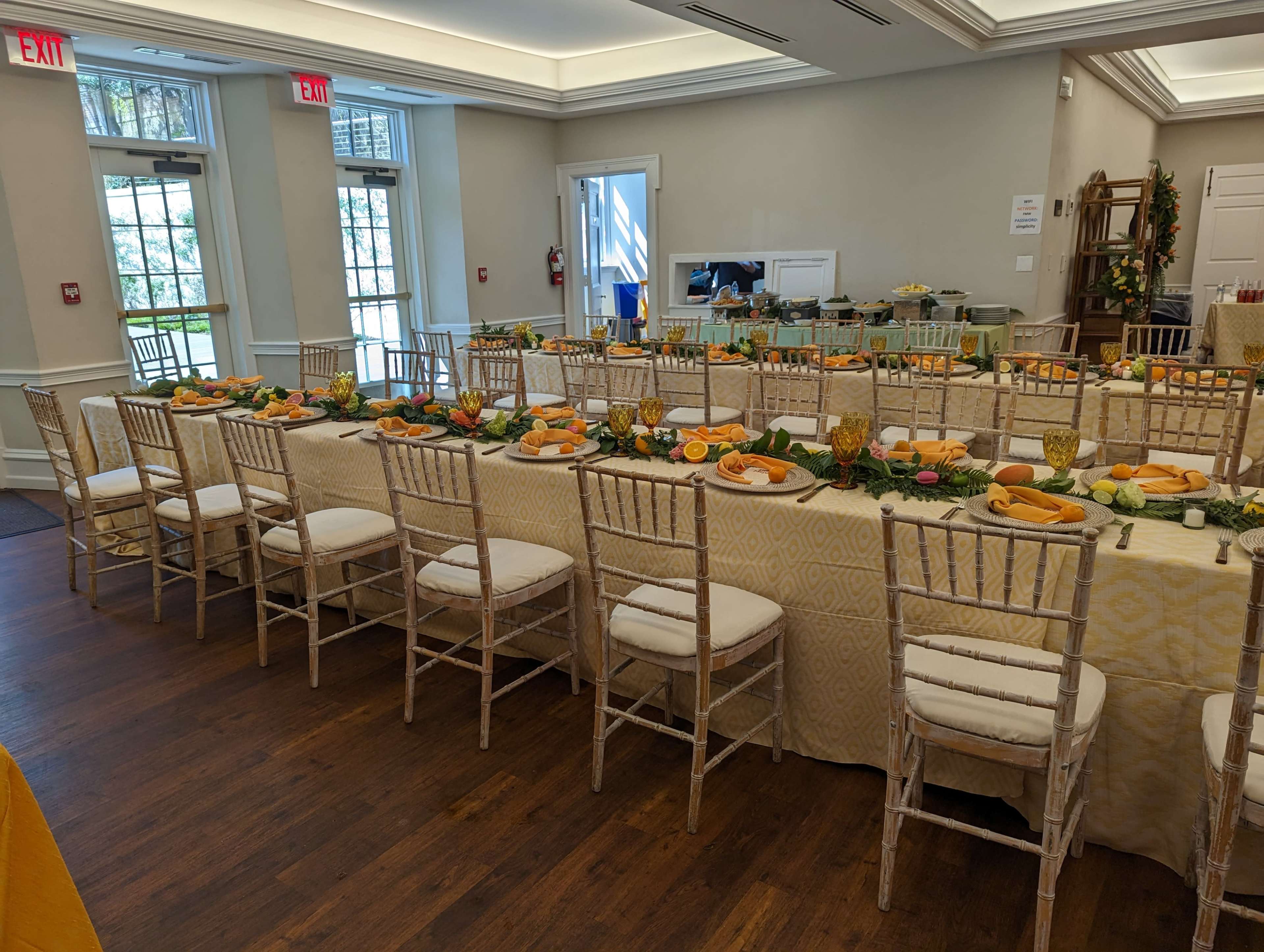 A dining setup features a long table adorned with orange fruits, greenery, and glassware in a well-lit room with large windows.