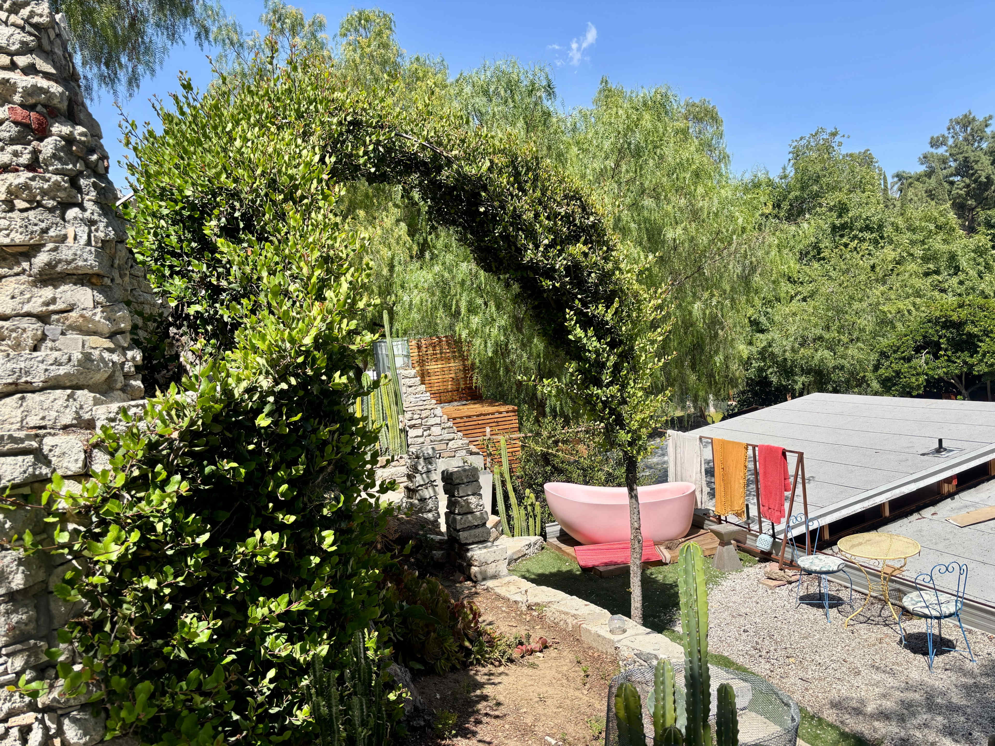 The scene features a garden with a stone arch covered in greenery, leading to a modern pink bathtub surrounded by trees and a rustic building with colorful panels.