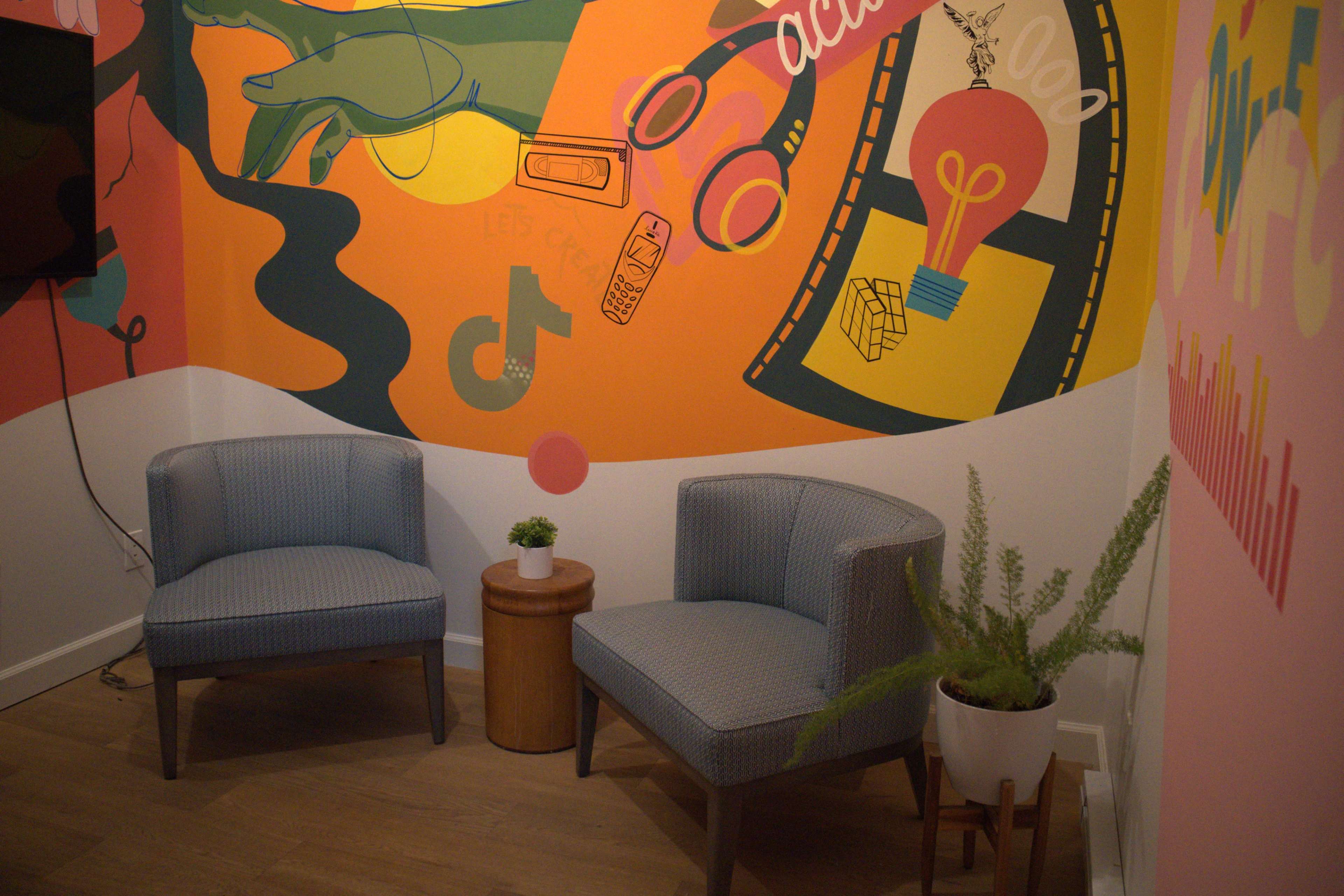 The image shows a brightly colored corner of a room featuring two gray armchairs, a small wooden table, and a potted plant against a mural backdrop.